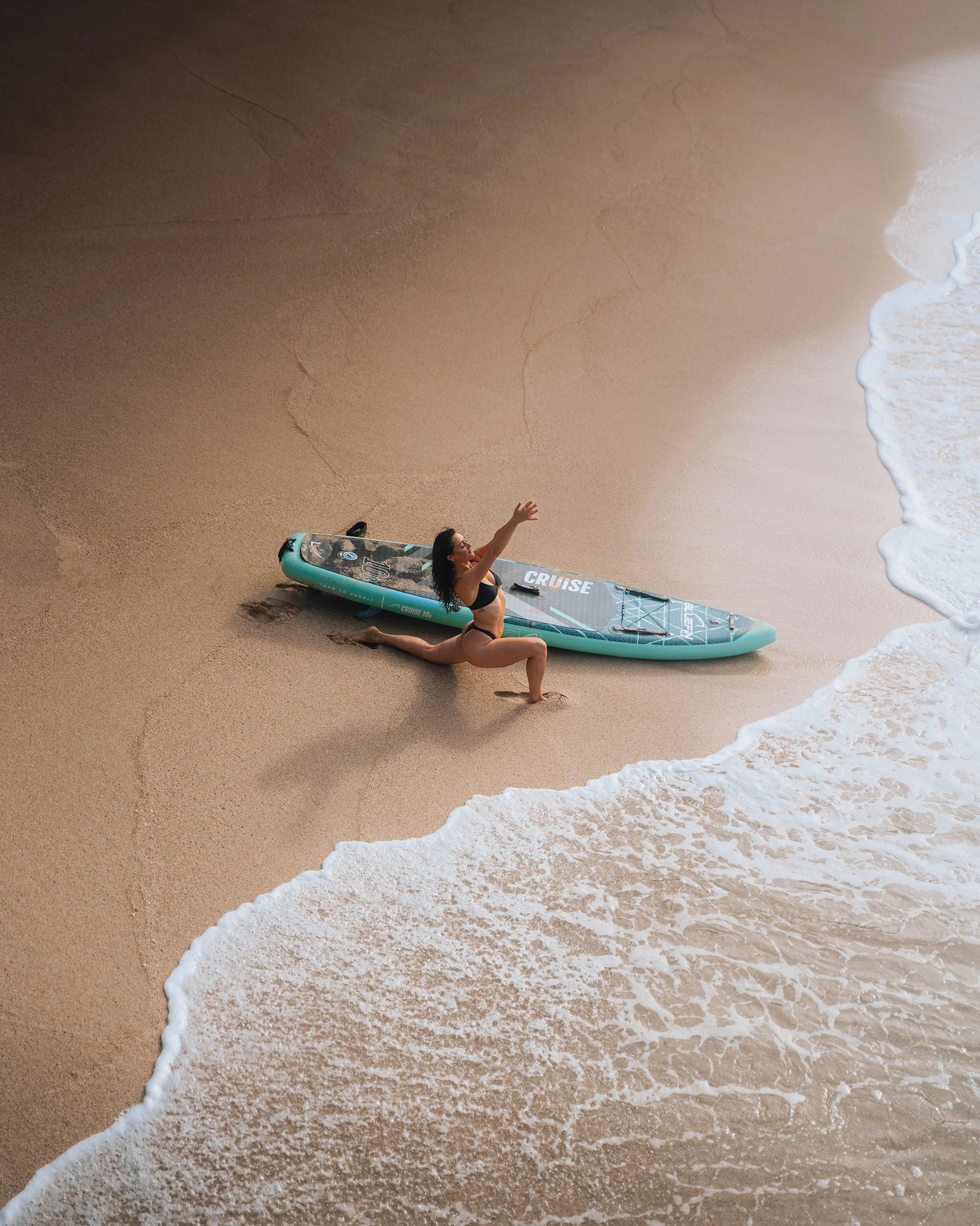 A woman in a black bikini practicing yoga on the sand next to a paddleboard at the beach, with ocean waves partially covering the shore.