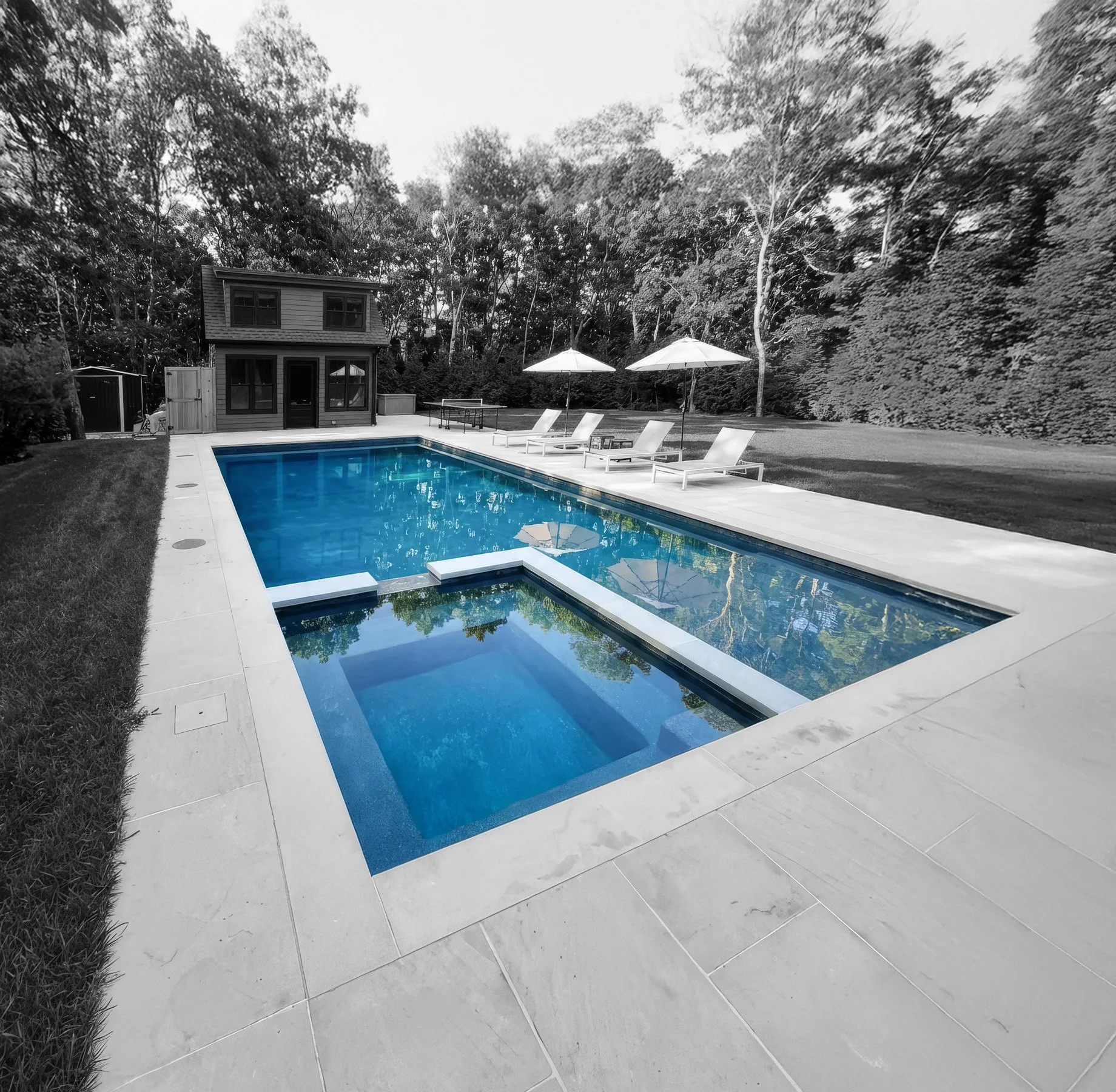 A backyard with a modern rectangular swimming pool with a spa section, surrounded by white stone paving, with three lounge chairs and two large umbrellas nearby. In the background, there is a small house or pool house and a wooded area with tall tree