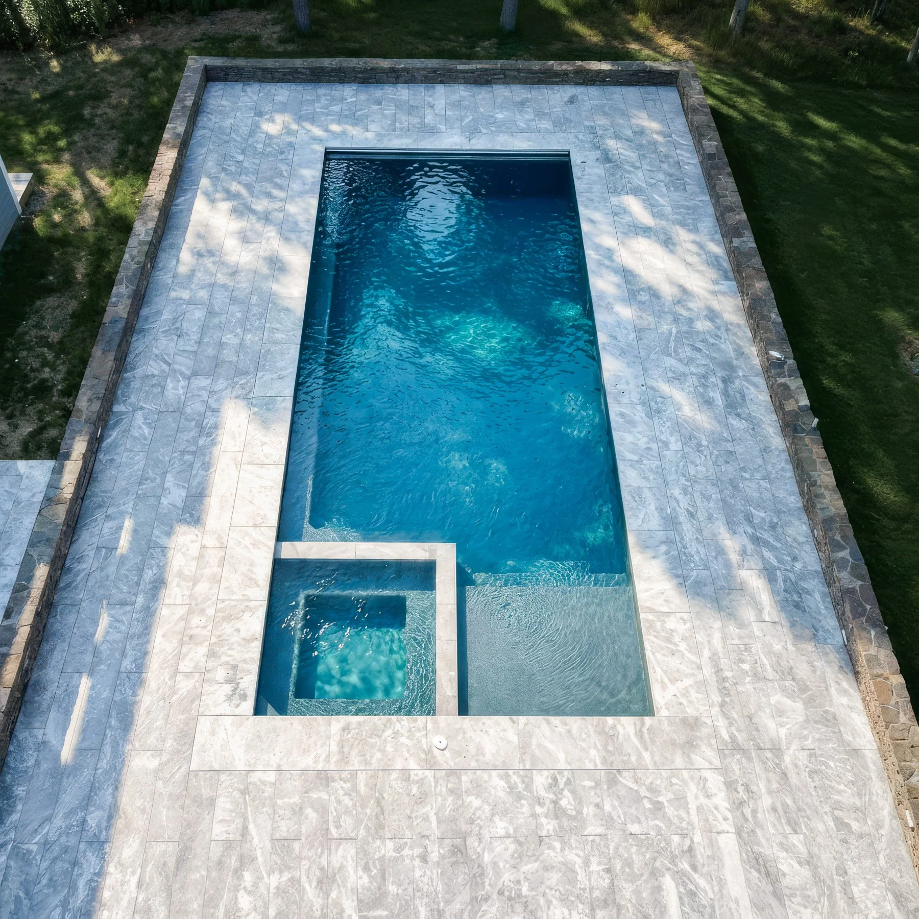 Aerial view of a rectangular in-ground swimming pool with a smaller attached spa, surrounded by a stone deck, in a backyard with grass and a few trees.