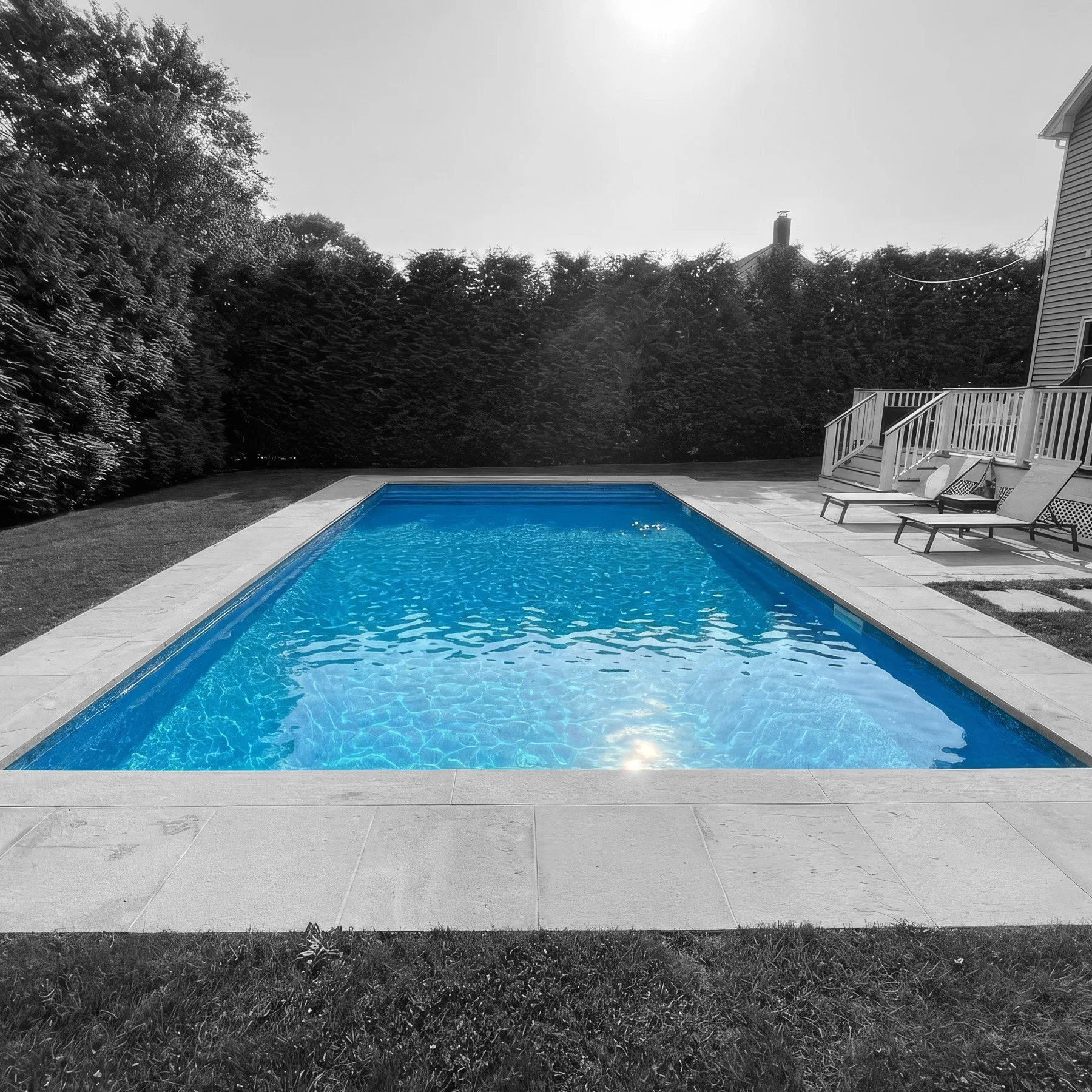 A swimming pool with blue water in a backyard, surrounded by a gray stone deck and a grassy lawn, with lounge chairs and a wooden deck with railings on the right side, and trees in the background.