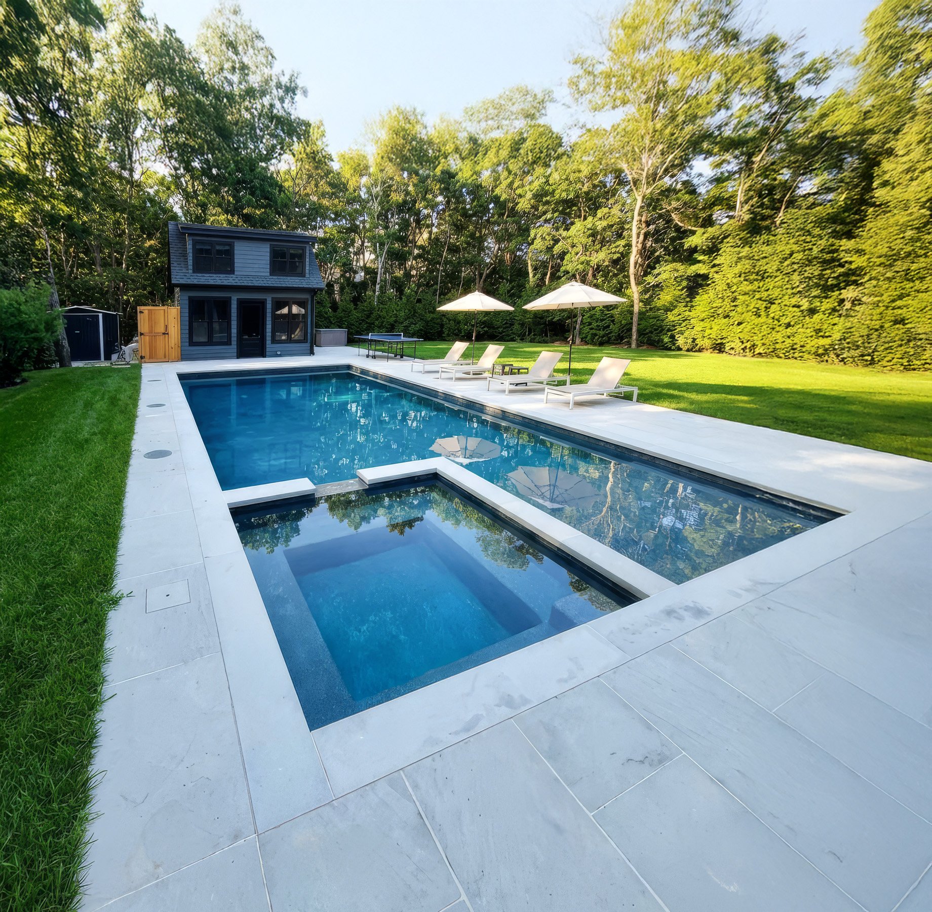Modern backyard with a rectangular swimming pool, pool house, lounge chairs under umbrellas, and lush green trees in the background.