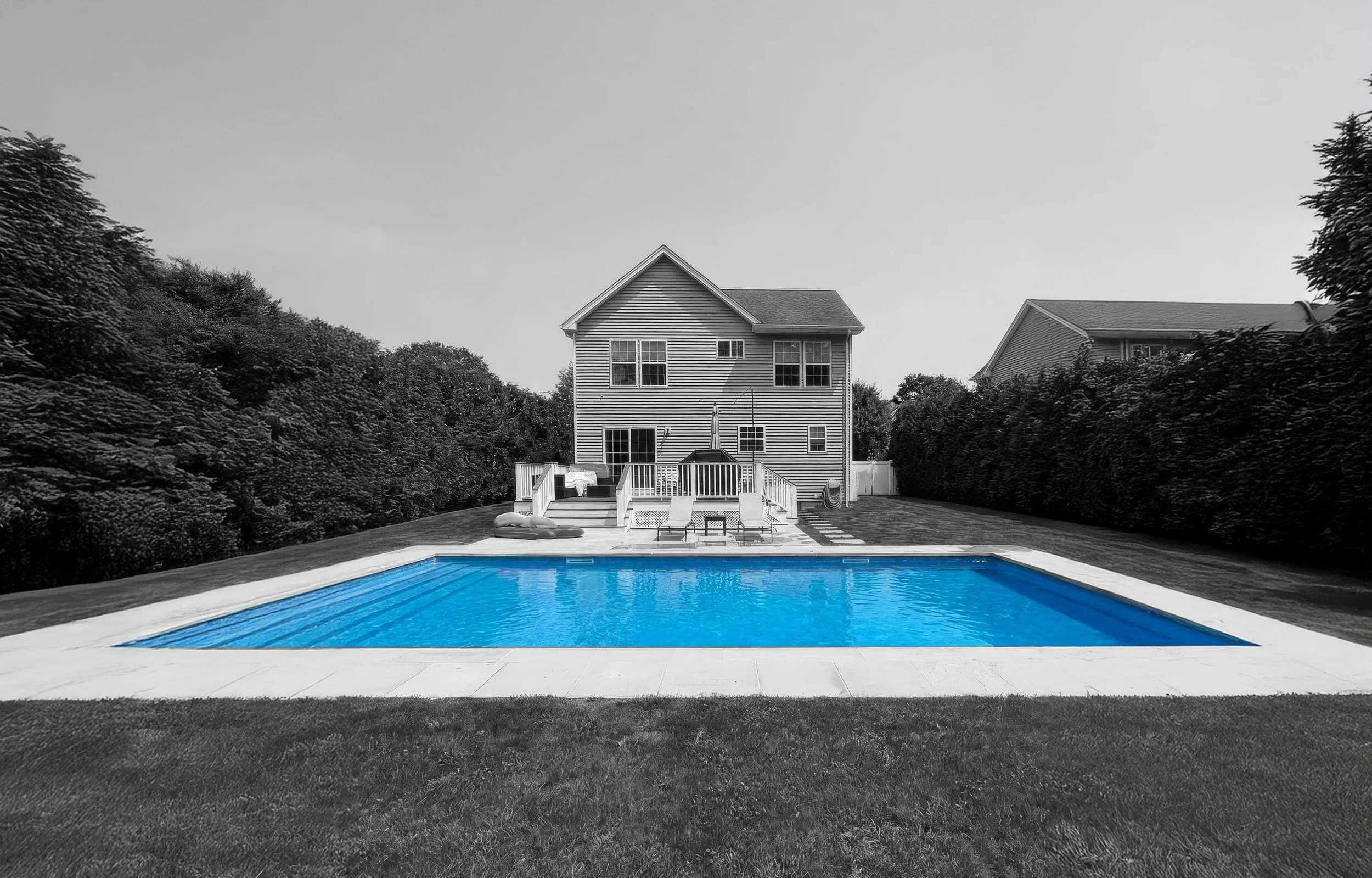 Backyard with a rectangular swimming pool filled with blue water, surrounded by a concrete deck, with a two-story house and lush hedges in the background.