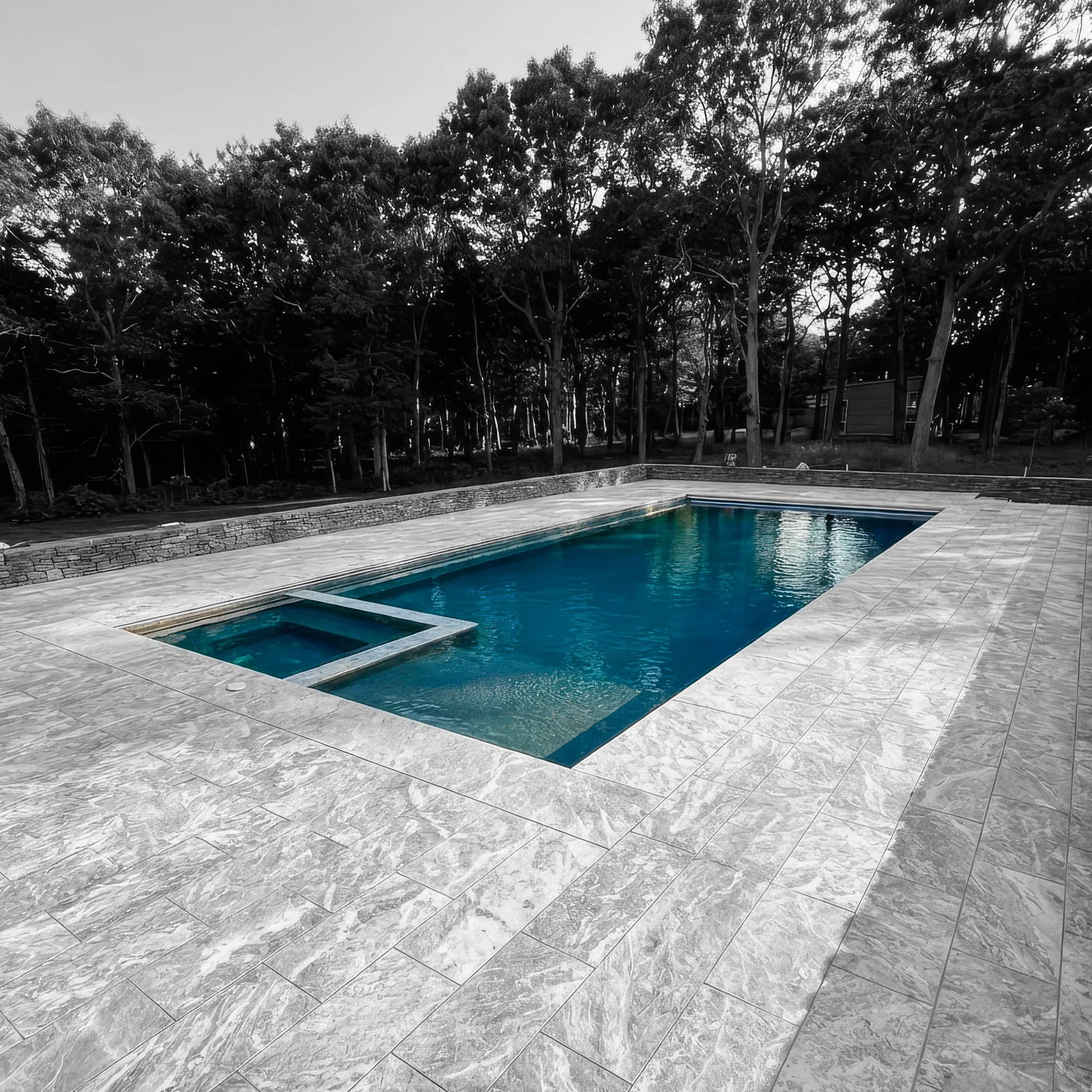 Color photo of a swimming pool with blue water, surrounded by a gray stone deck, with a wooded area in the background.