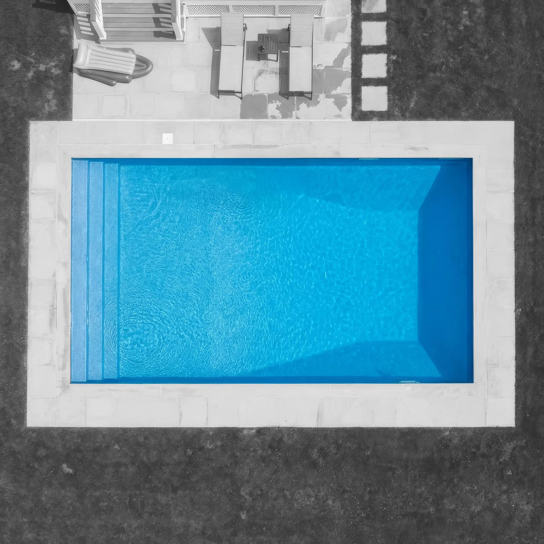  aerial view of a rectangular swimming pool filled with blue water, surrounded by a white stone deck and outdoor furniture, with a mix of grass and concrete areas around it.
