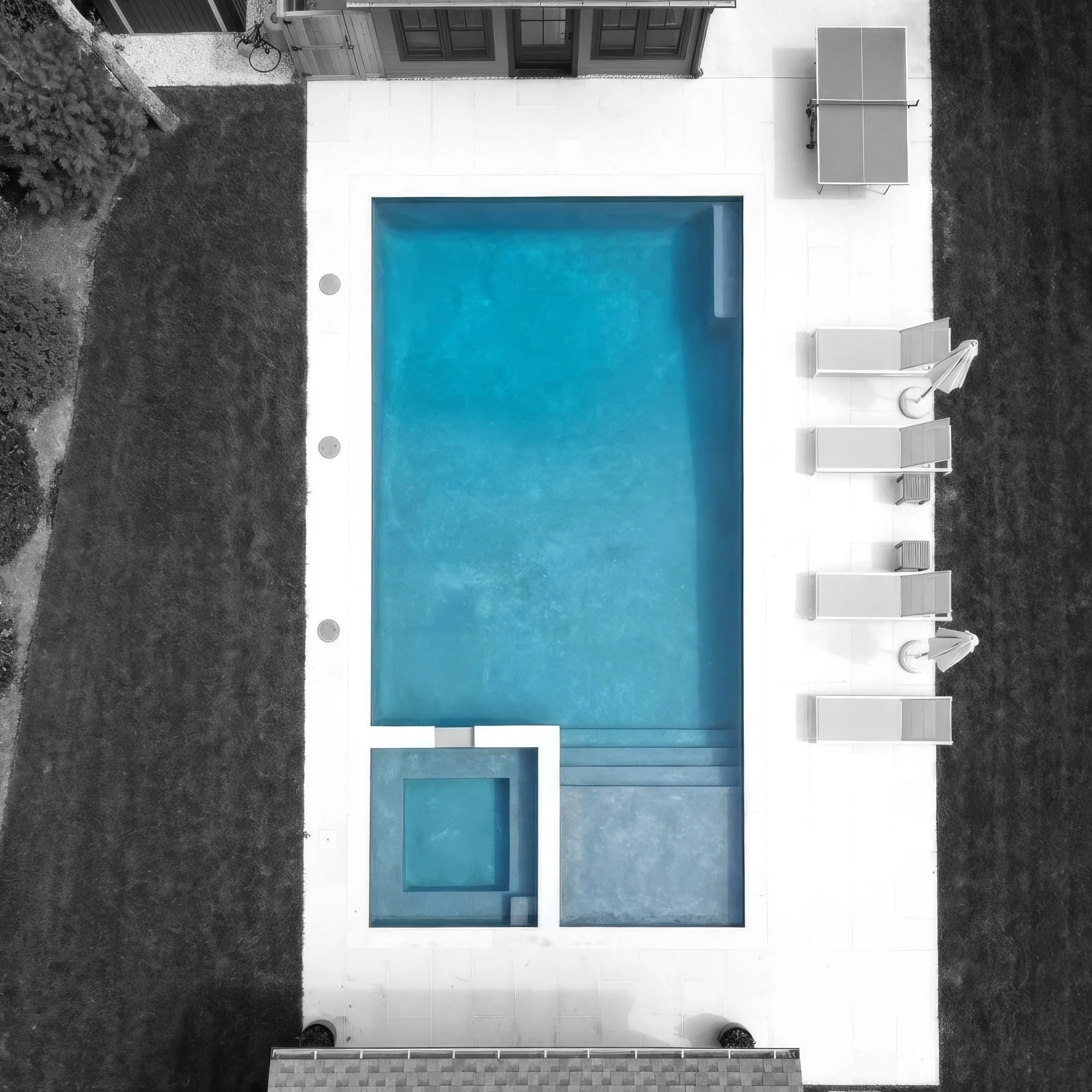 A backyard with an in-ground swimming pool surrounded by a white patio. The pool has a smaller hot tub attached at one end, and several white lounge chairs and patio furniture are visible on the patio. There is grass and shrubbery around the patio ar