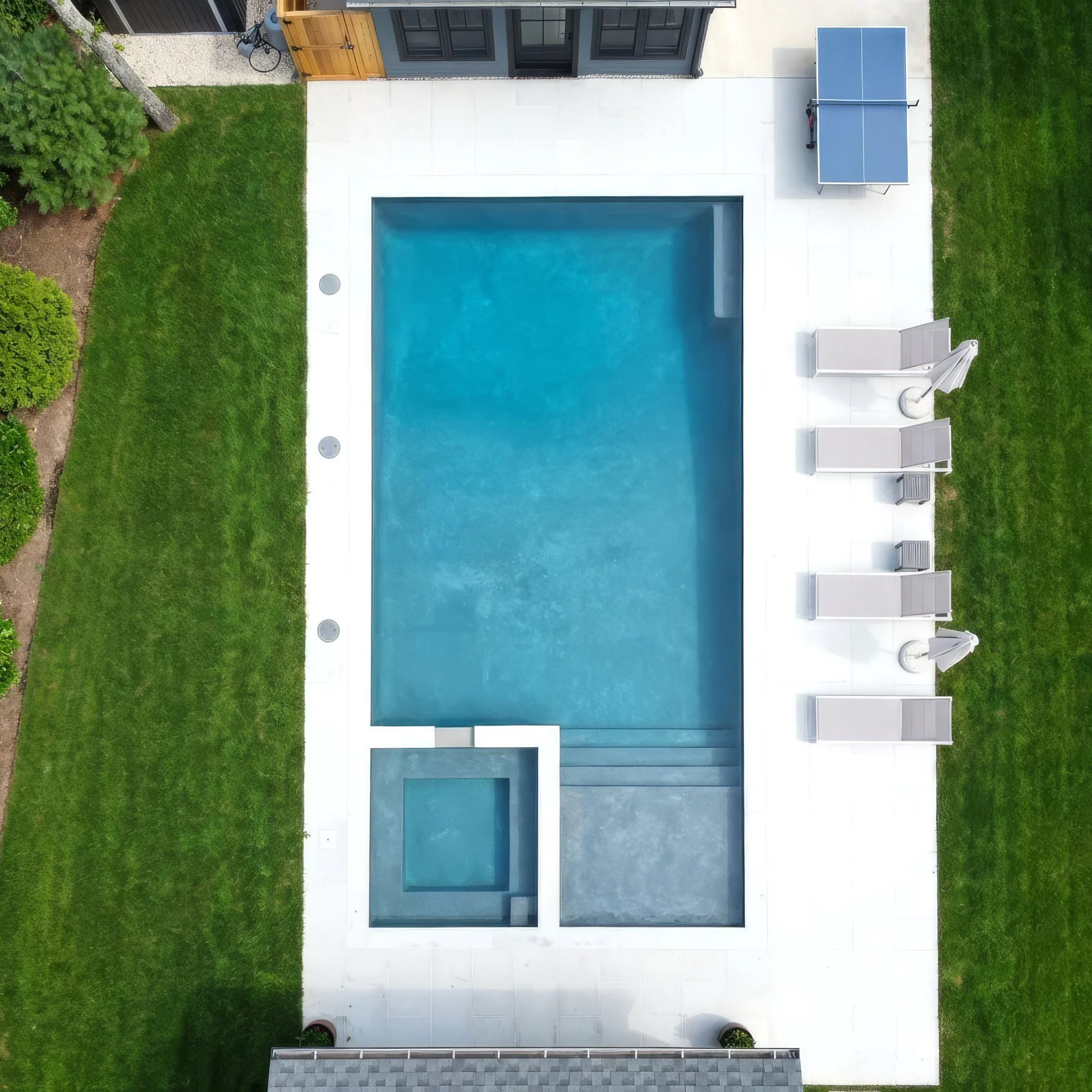 An aerial view of a backyard with a rectangular swimming pool surrounded by white paving. There are six lounge chairs with umbrellas along the right side of the pool, a small table, and a wiffle ball set on the lawn. The yard has green grass, trees, 