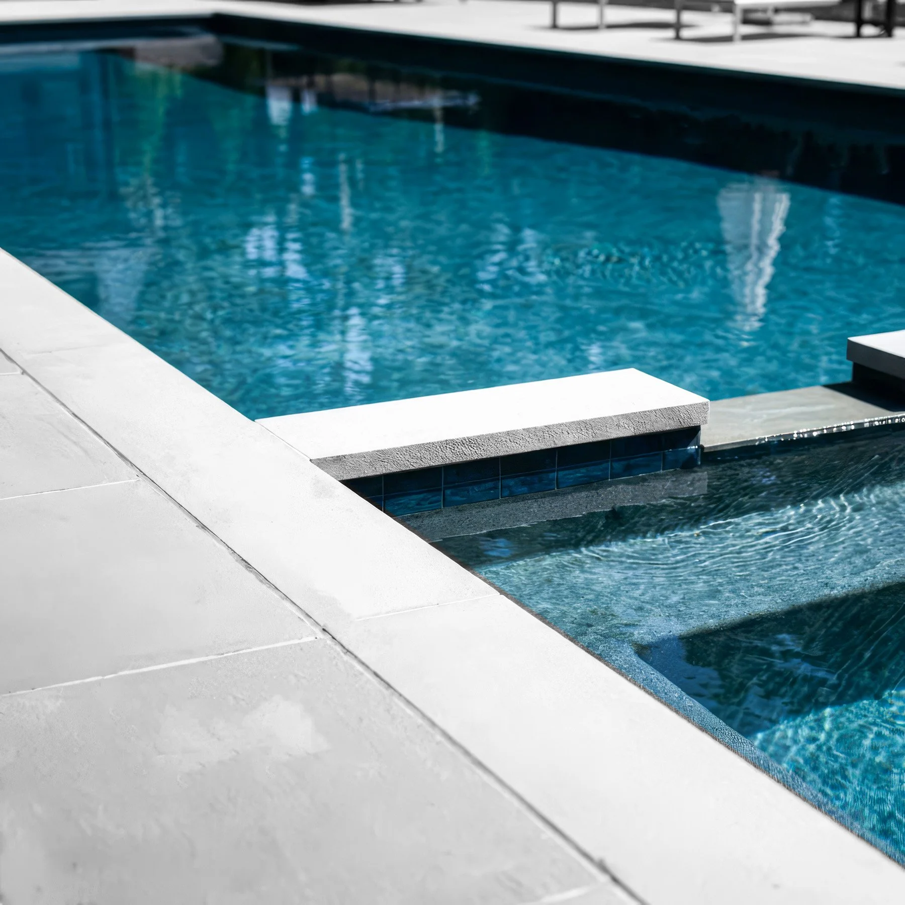 Close-up of a modern swimming pool edge with white tiles and a blue tiled water channel, with a clear view of the pool water in the background.