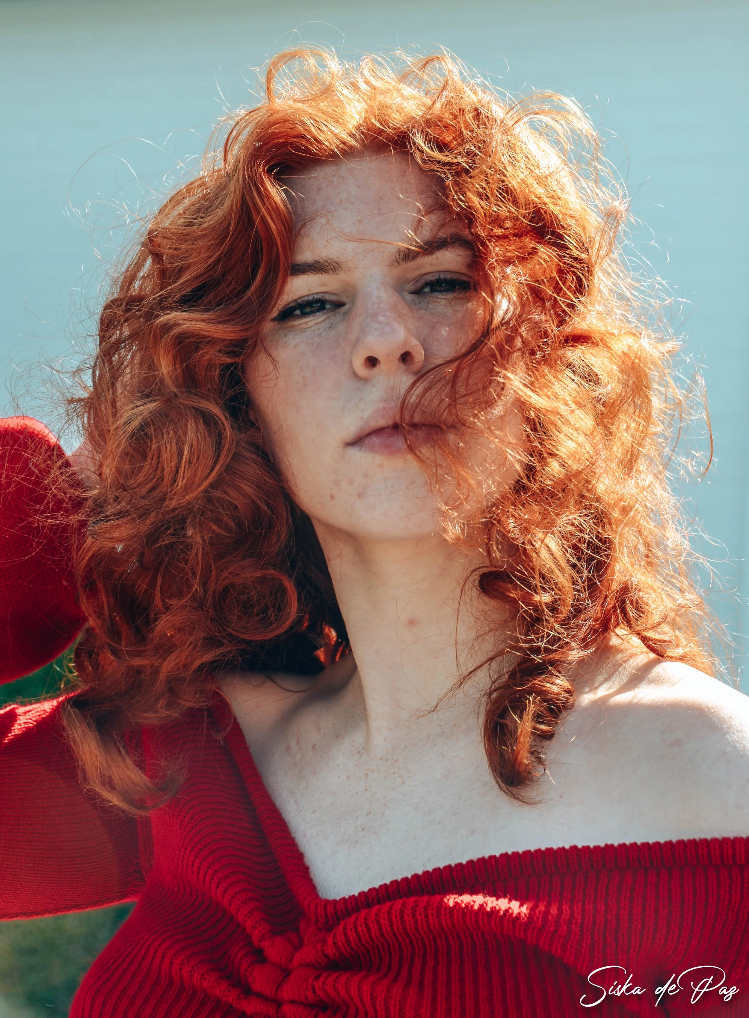 Close-up of a young woman with red, curly hair, wearing a red off-shoulder top, standing outdoors with a light blue sky background.