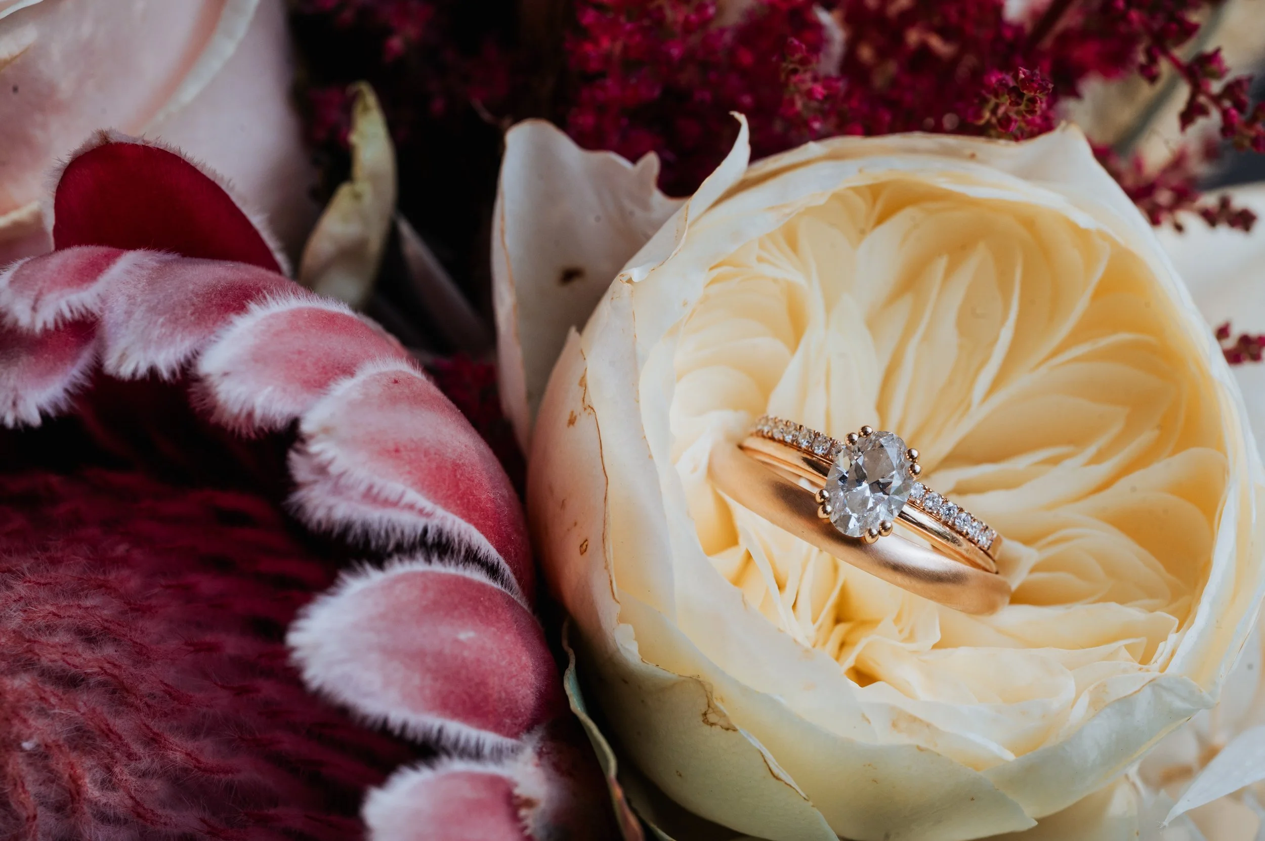 A diamond engagement ring with a large oval-cut diamond in a rose gold band, placed inside a cream-colored rose blossom, surrounded by various flowers.
