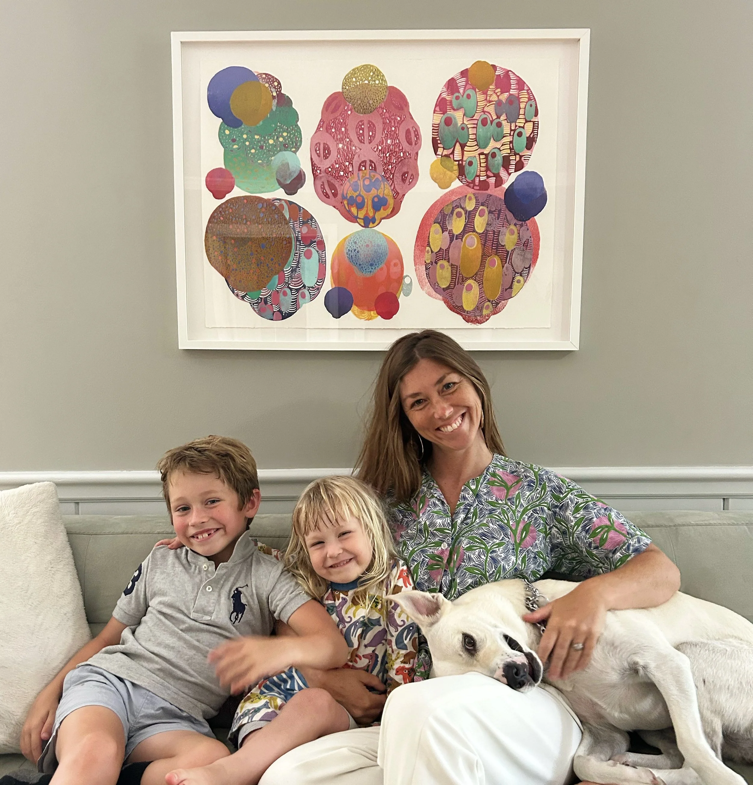 A woman with two children and a dog sitting on a sofa in a living room with a colorful abstract art piece hanging on the wall behind them.