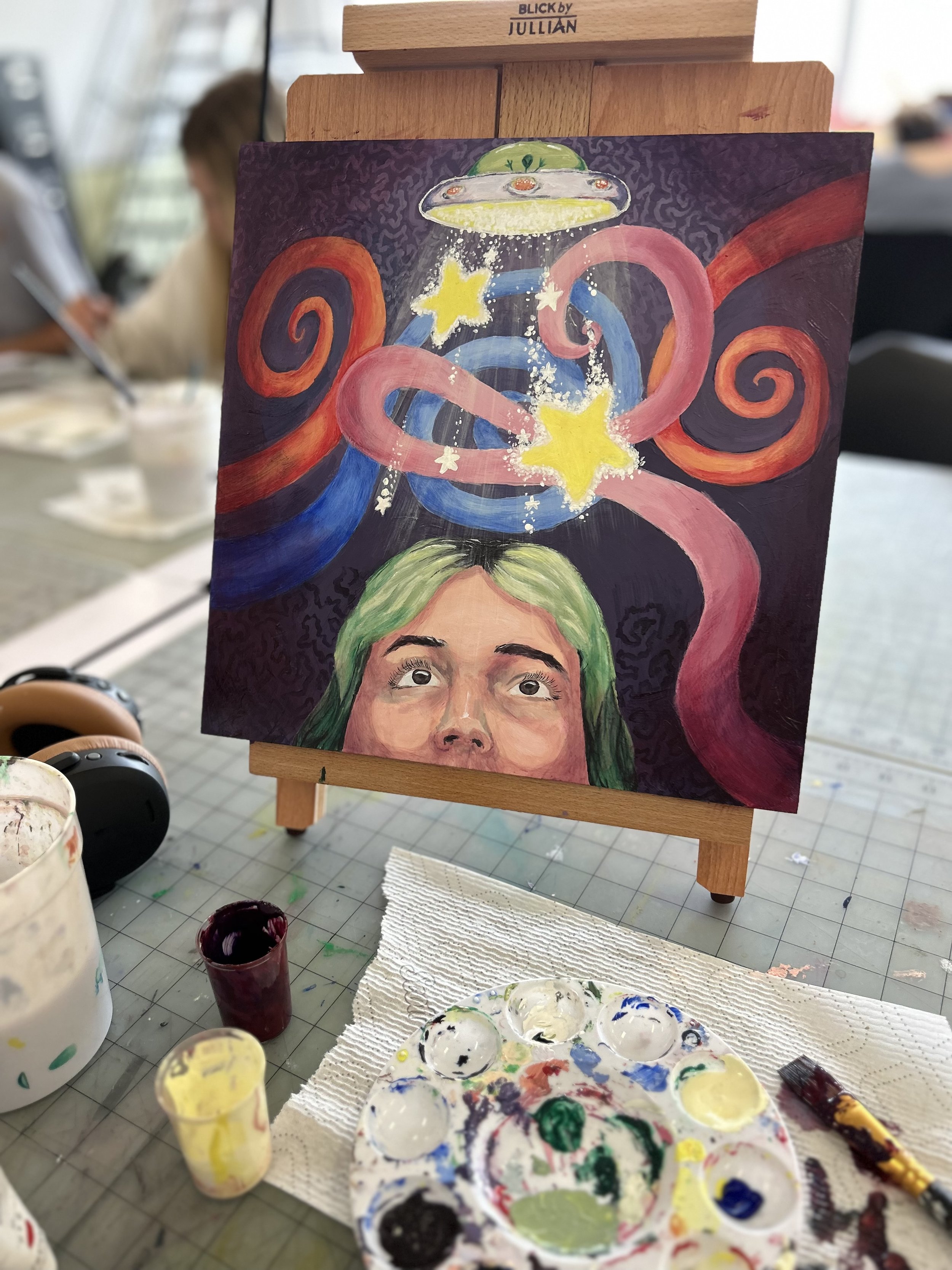 A colorful painting of a woman with green hair, looking up at a flying saucer emitting yellow stars and pink, blue, and red swirling shapes, on an easel at an art studio.