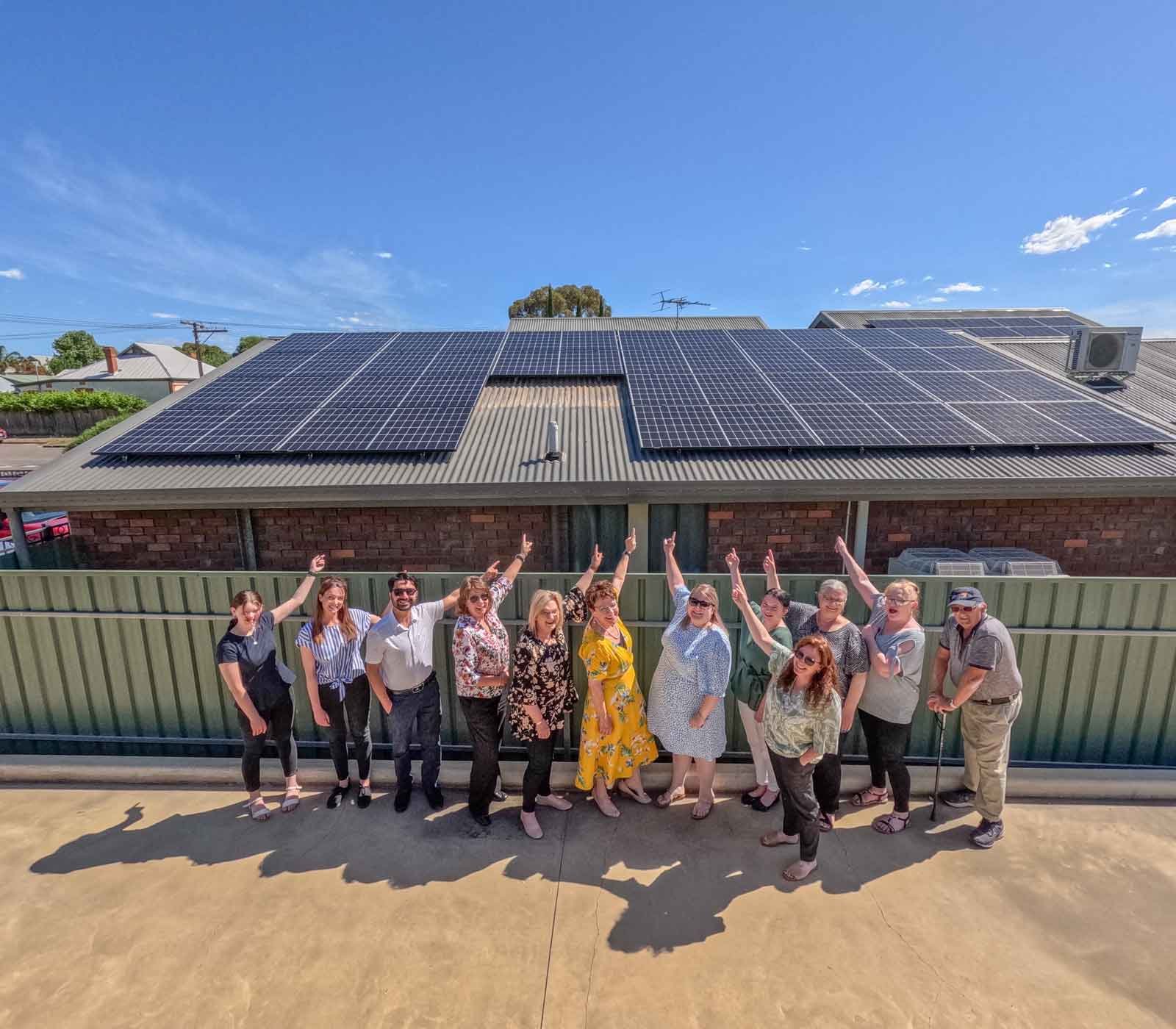 Group of 11 people standing in front of a brick building with a large solar panel array on the roof, raising their arms in celebration under a clear blue sky.