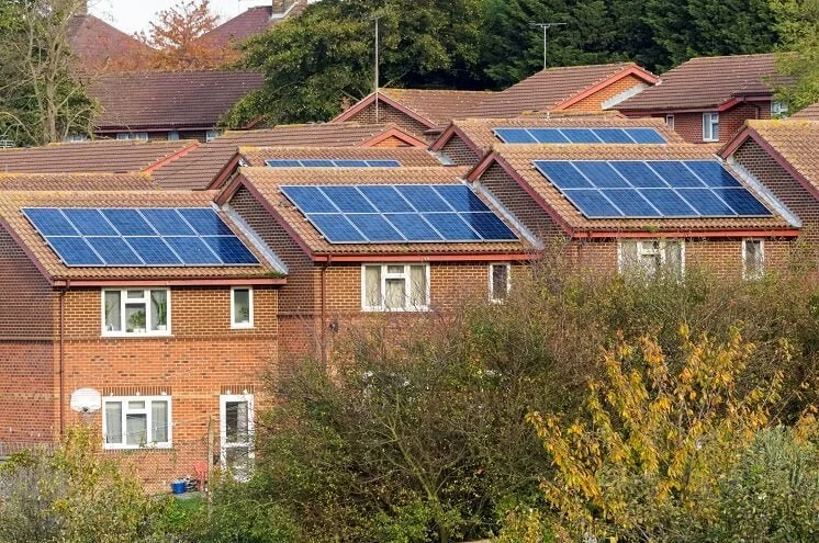 Row of brick semi-detached houses with tiled roofs, each fitted with arrays of blue solar panels on the south-facing slopes, surrounded by greenery.