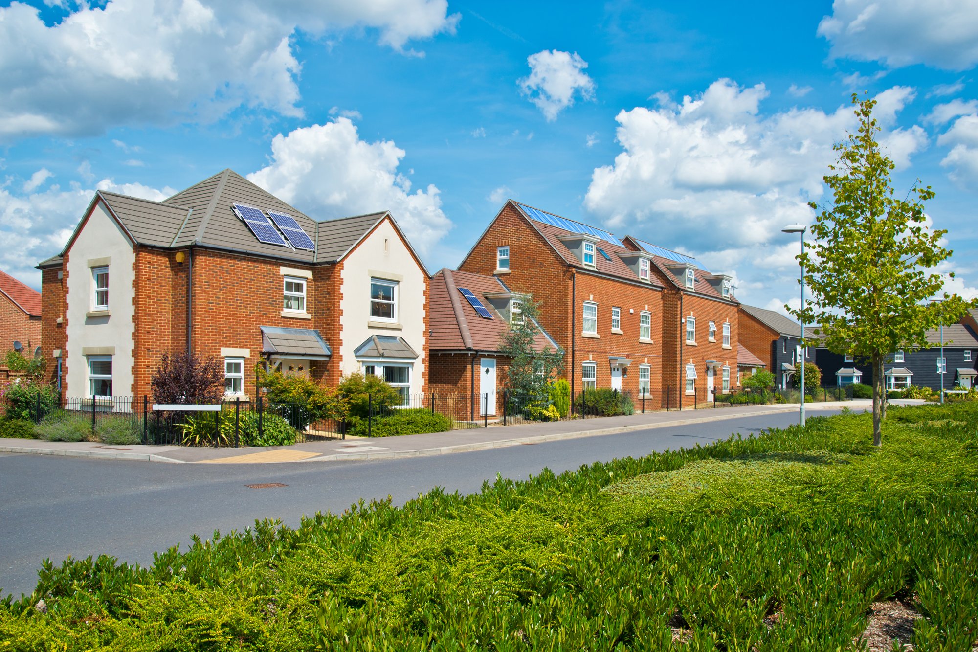 Residential neighborhood with modern brick houses featuring solar panels, green lawns, trees, and a clear blue sky.