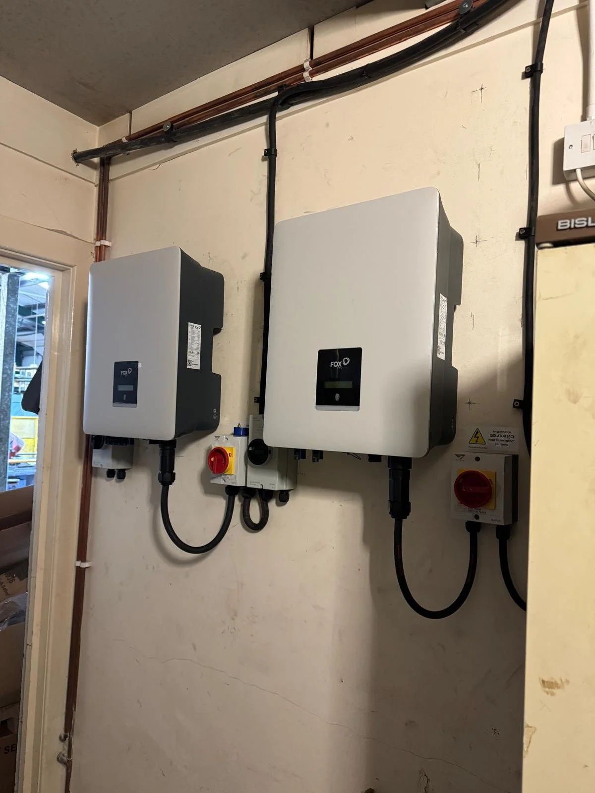 Two Fox ESS inverter units wall-mounted side by side with isolator switches and cabling in a utility room.