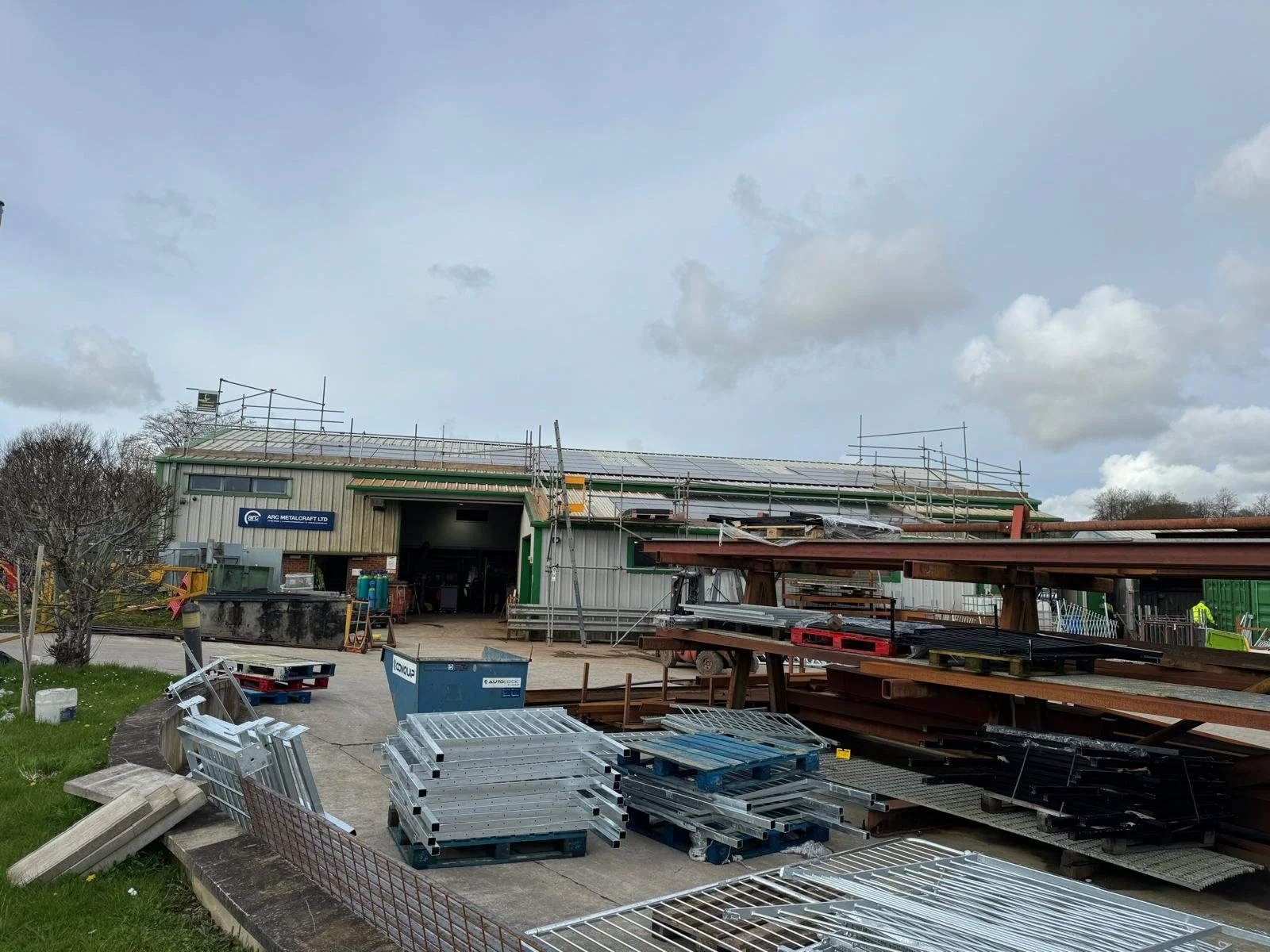 Industrial workshop yard with scaffolding on the roof and stacks of steel beams and metal panels in the foreground.