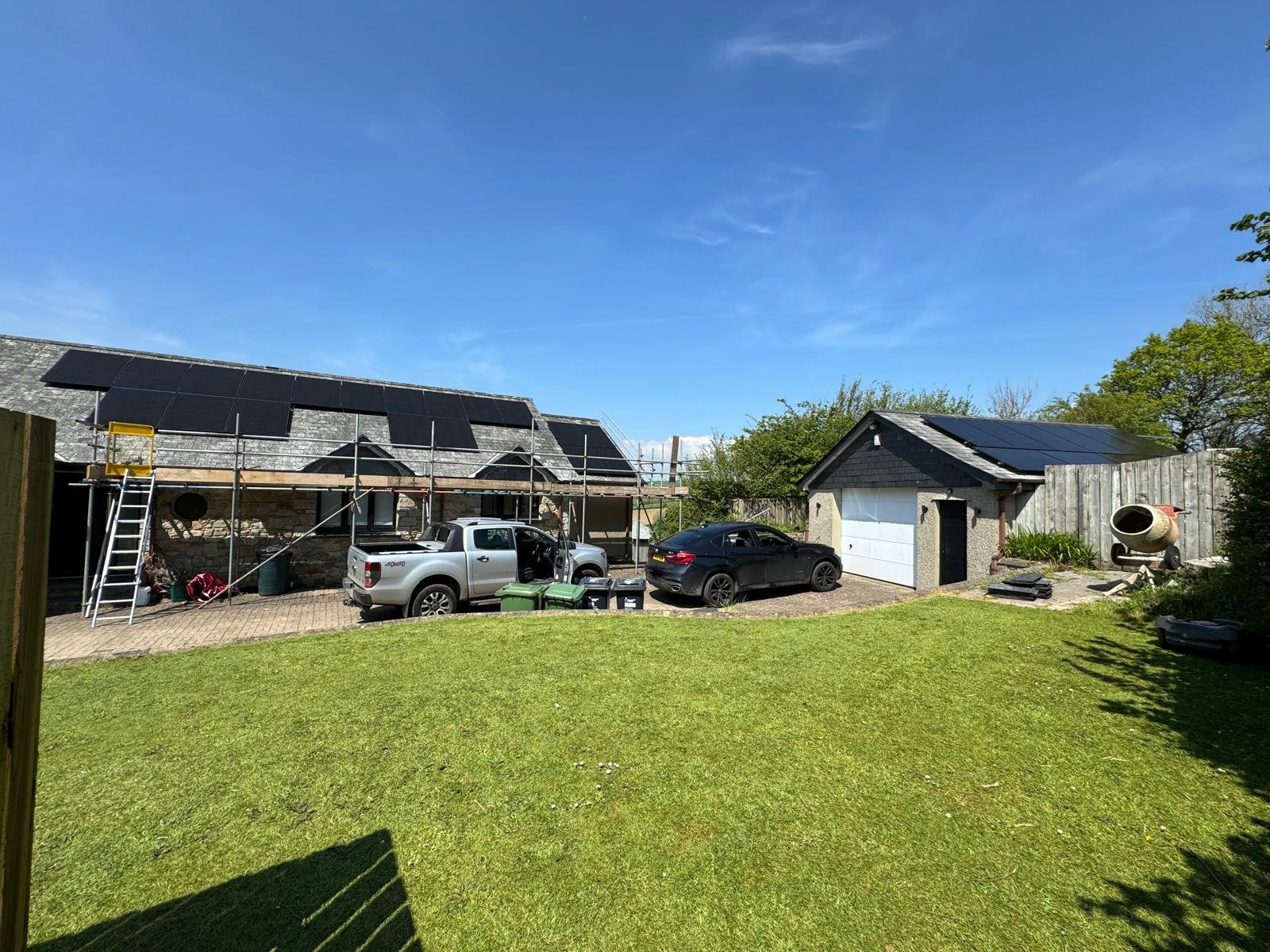 Residential backyard with a house under renovation, scaffolding on the left side, solar panels on the roof, two cars parked, a cement mixer on the right, and a grassy yard in the foreground under a clear blue sky.