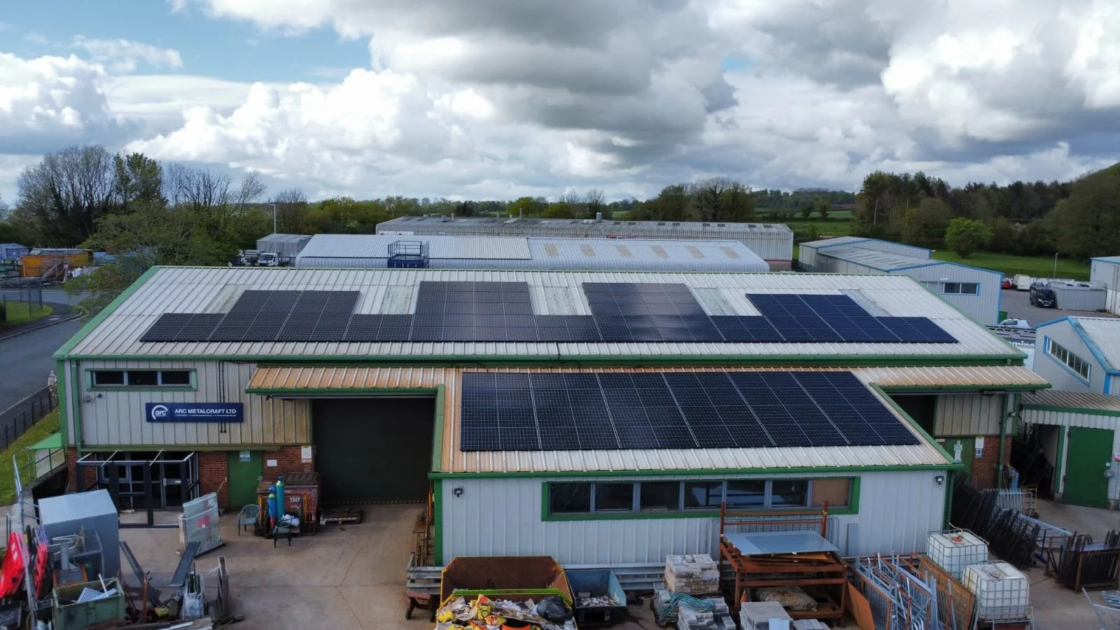 Industrial building with solar panels on the roof, surrounded by trees and clouds in the sky.