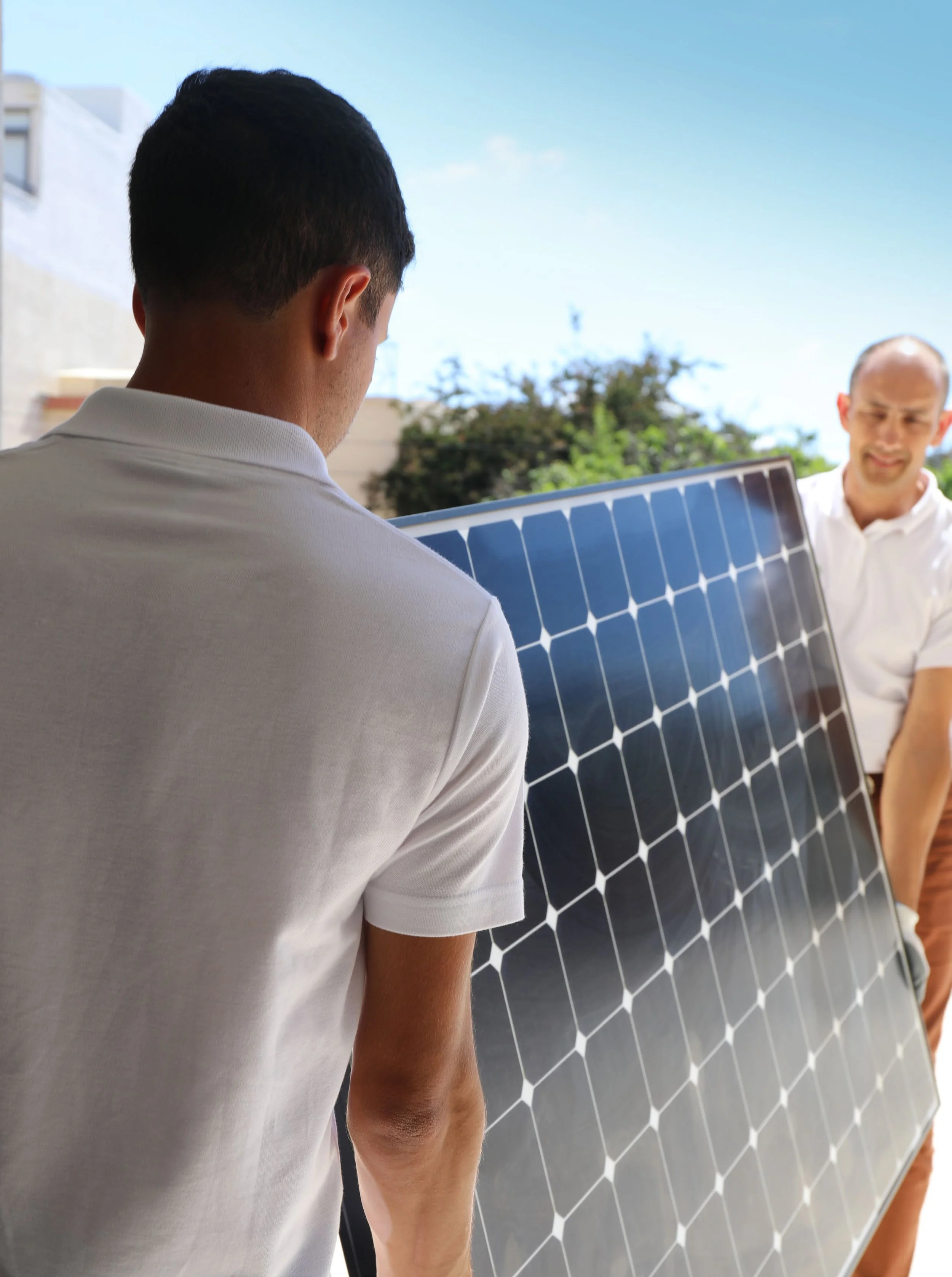 Two installers in white shirts carrying a large blue polycrystalline solar panel outdoors on a sunny day.