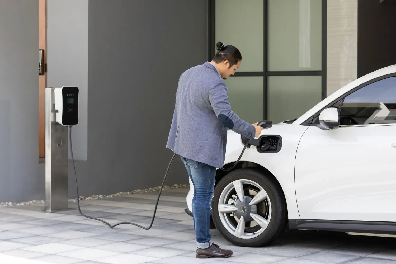 Man with man bun plugging a charging cable into a white electric SUV at a wall-mounted EV charger outside a modern building.