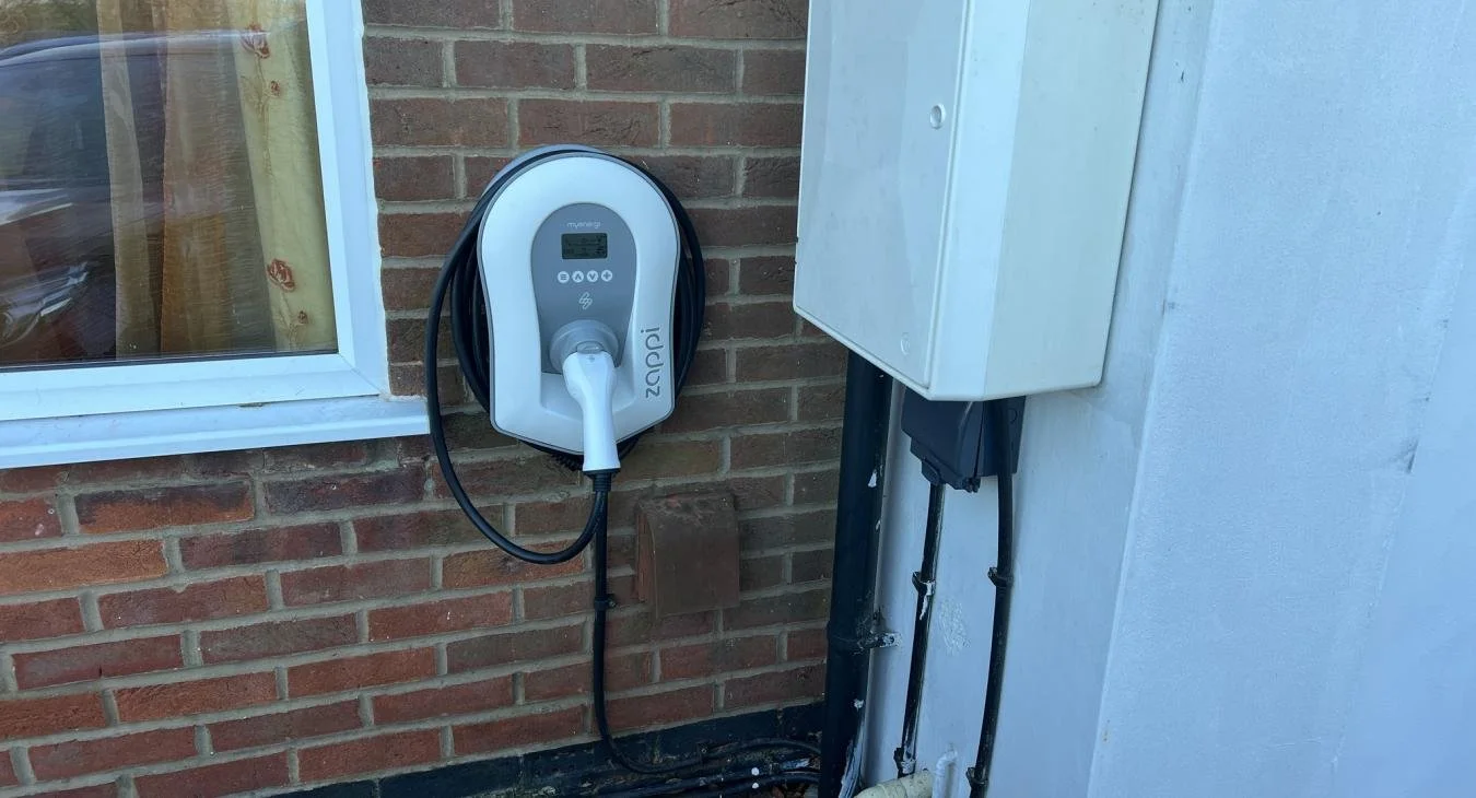 Wall-mounted myenergi Zappi EV charger with tethered cable and digital display, installed on a brick wall next to a window and white utility box.