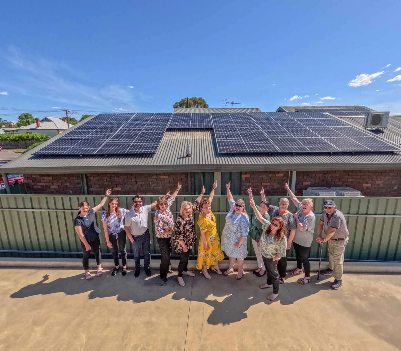 Group of 11 people standing in front of a brick building with a large solar panel array on the roof, raising their arms in celebration under a clear blue sky.