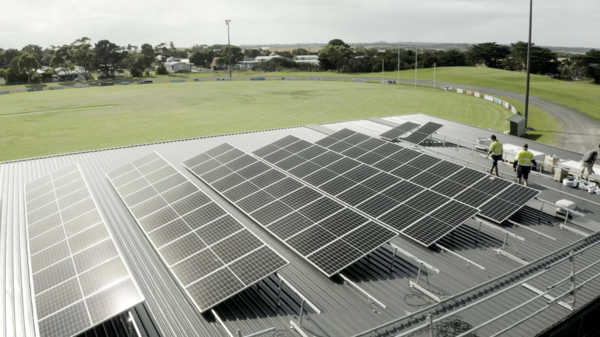 Aerial view of large solar panel arrays on the roof of a sports facility building, with two workers in hi-vis on the roof; green sports field in the background.