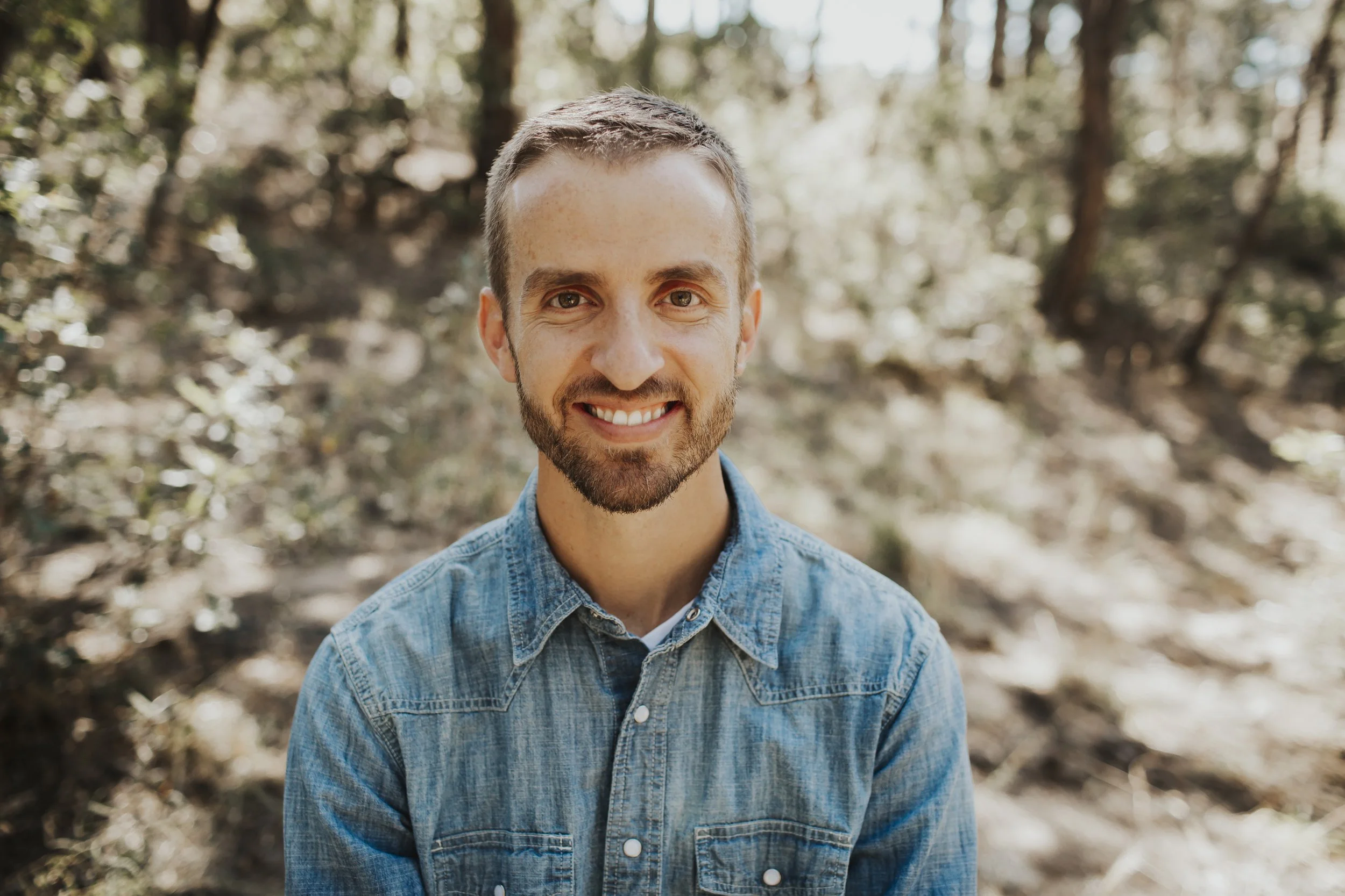 A smiling man with short brown hair and a beard, wearing a denim shirt, standing outdoors in a wooded area with trees and sunlight in the background.