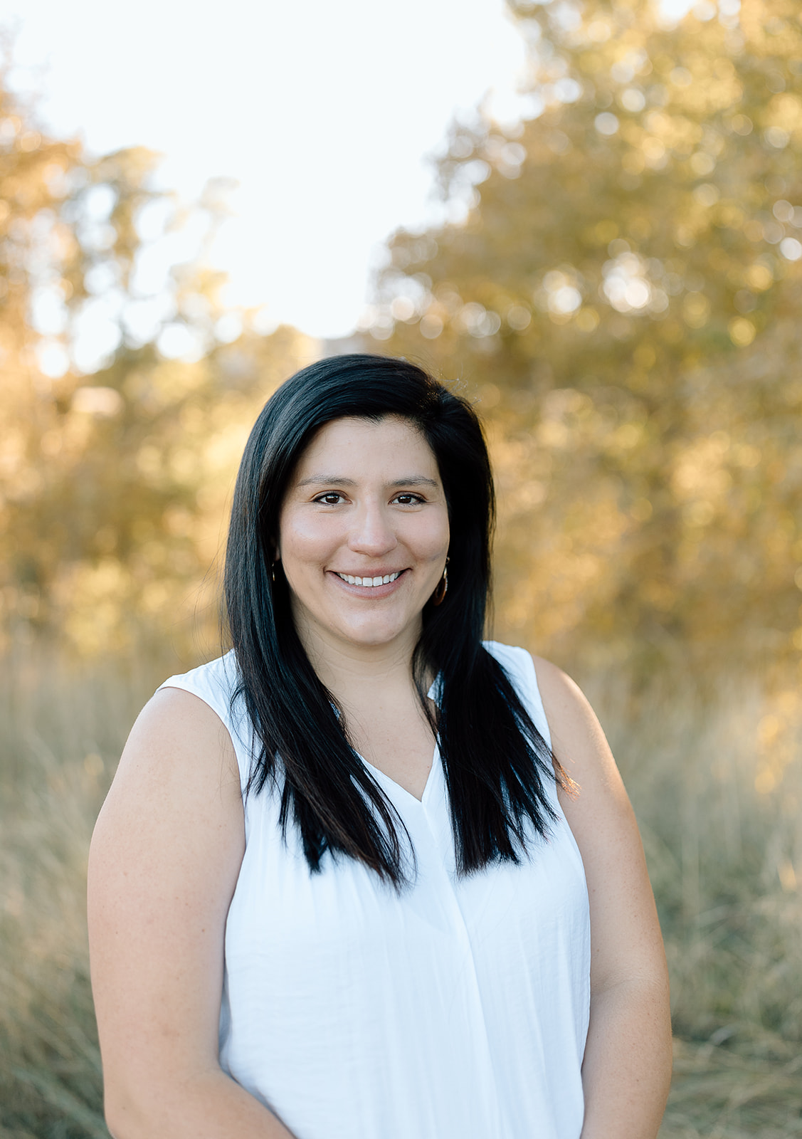 A woman with black hair smiling outdoors with autumn trees and sunlight in the background.