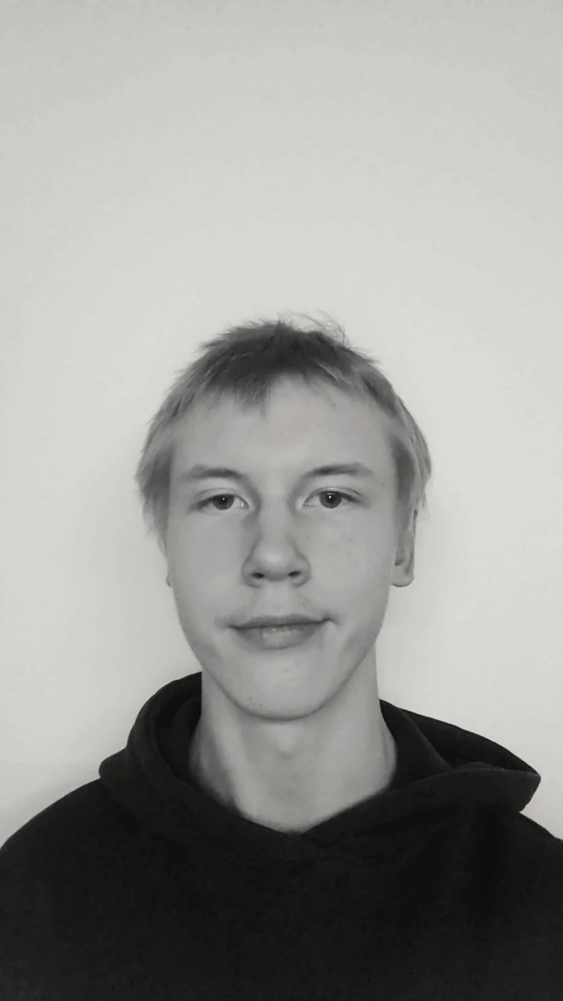 Young man with short hair wearing a black hoodie, looking at the camera, with a plain light-colored background.