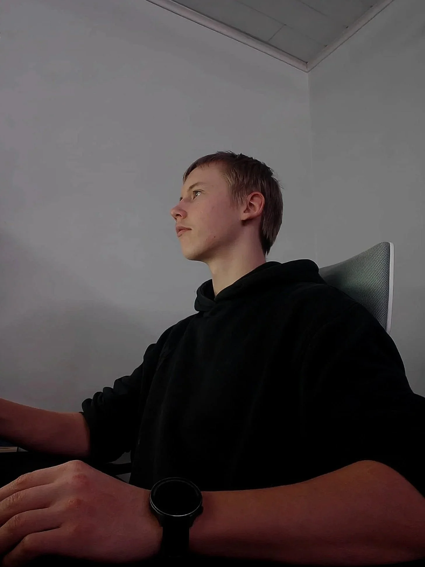 Side profile of a young man sitting at a desk, wearing a black hoodie and a black smartwatch, against a plain light grey wall.