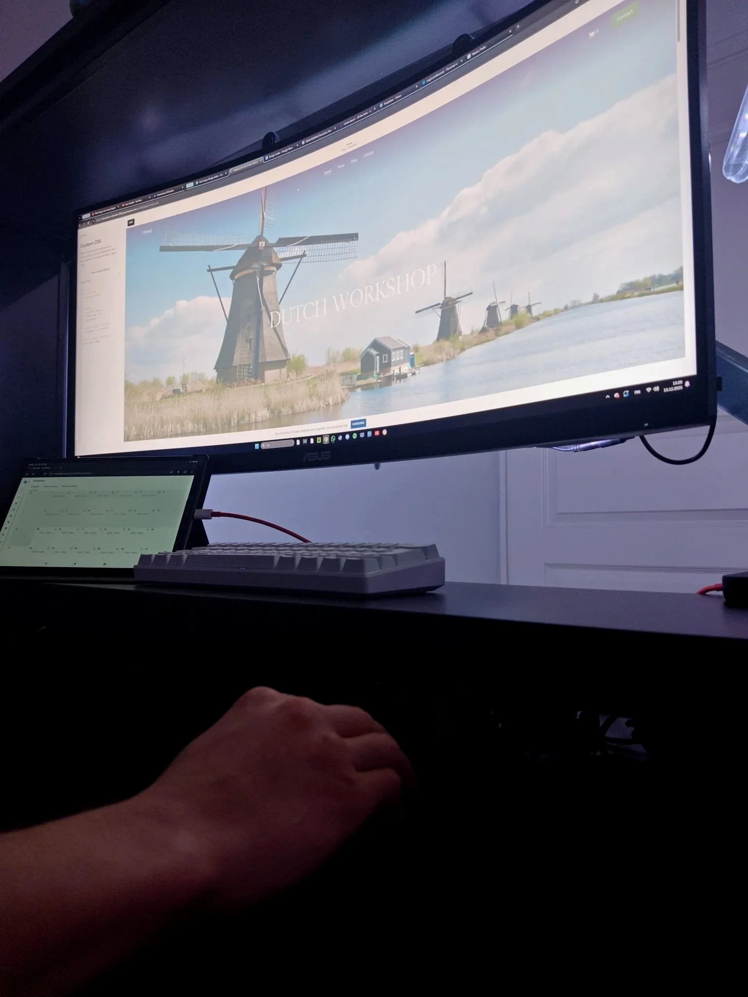 A computer monitor displays a website with the text "Dutch Workshop" and an image of windmills along a waterway. A person is using a keyboard and mouse in front of the monitor. A tablet is visible on the desk, showing some code or text.