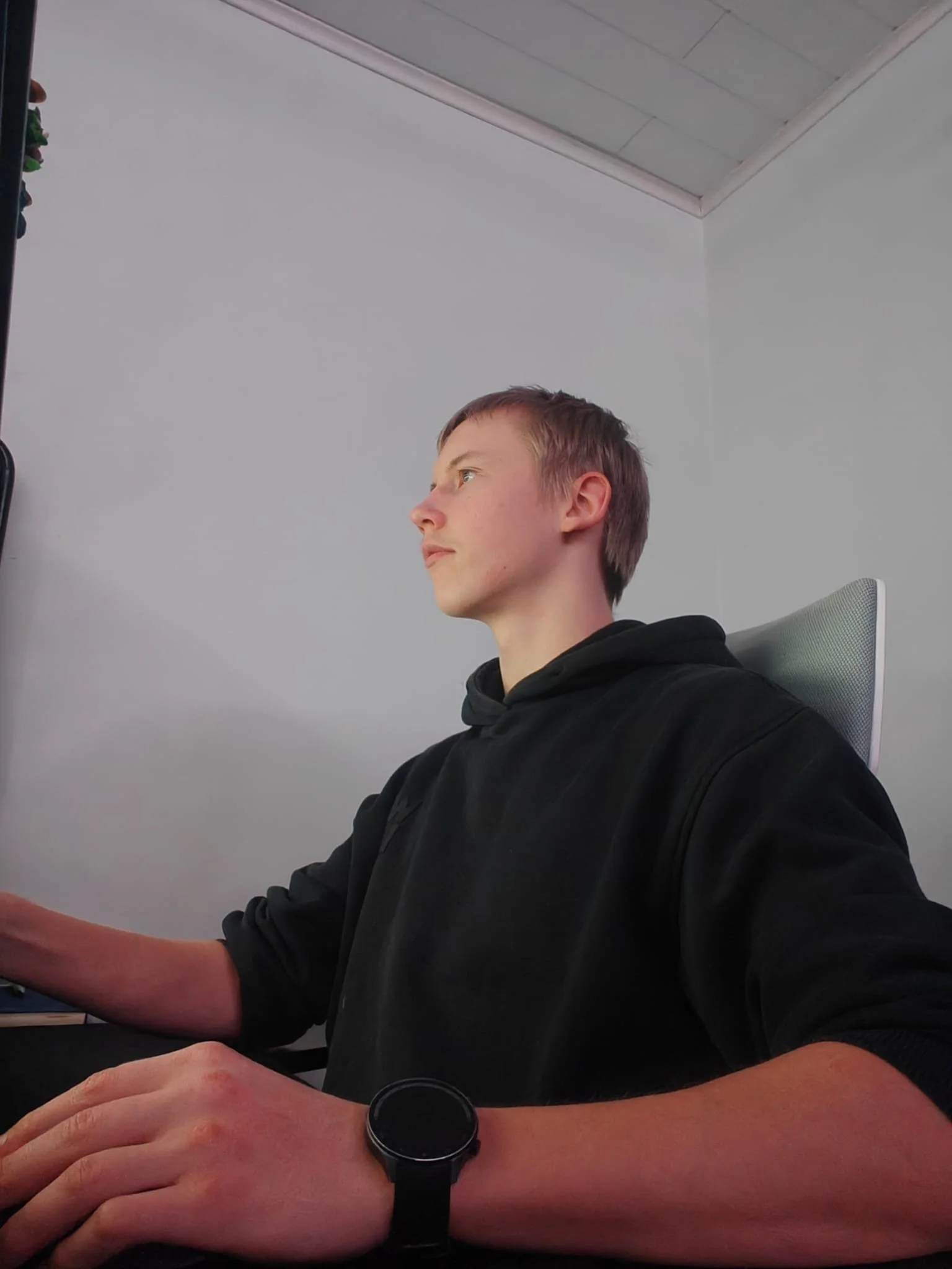 A young man with short hair sitting at a desk, wearing a black hoodie and a black wristwatch, focused on a computer screen in a plain room with white walls.