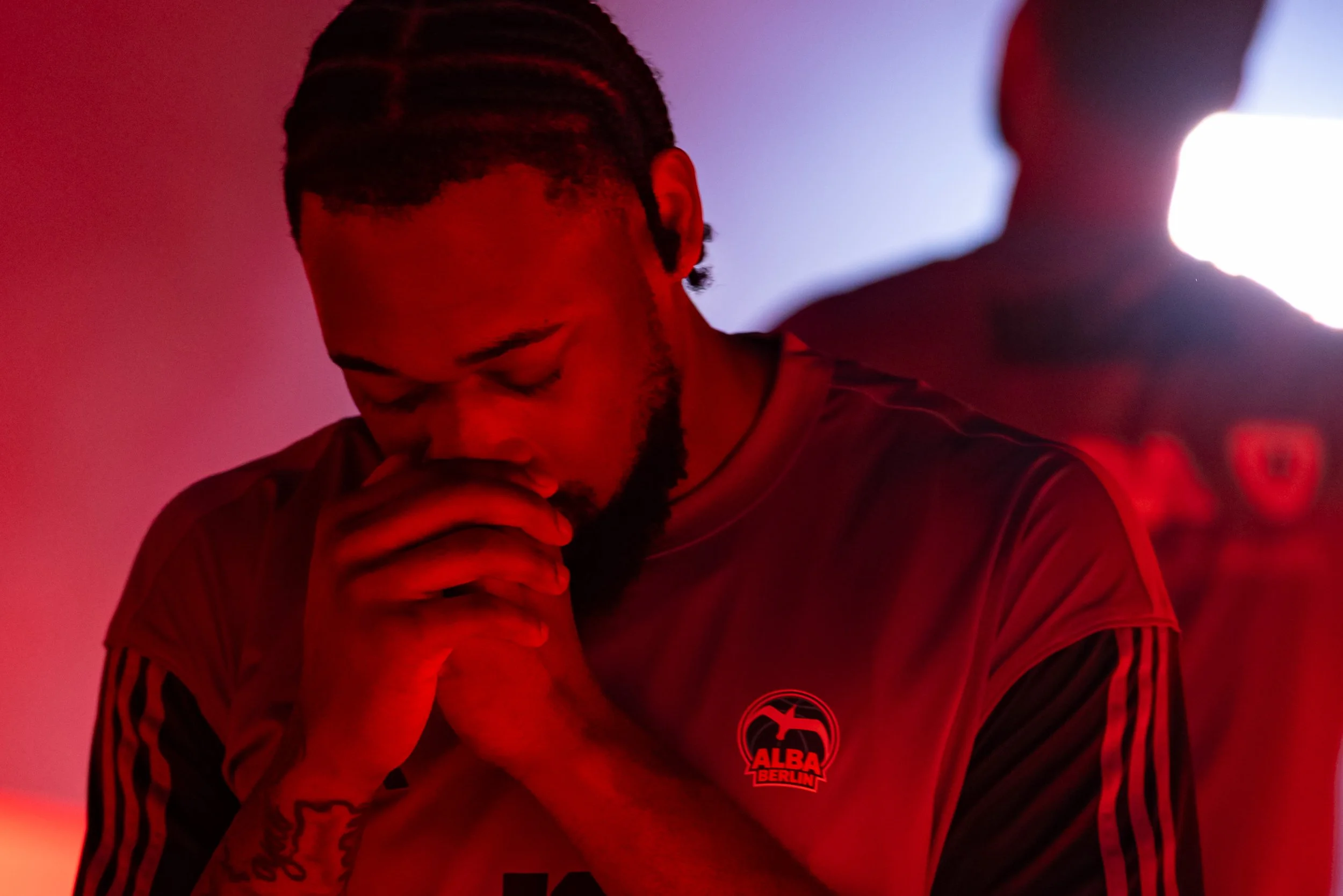 Ein junger Mann mit dunklen Haaren und Bart, trägt ein schwarzes Adidas-Trikot mit roten Streifen und einem ALBA Berlin Logo, sitzt in einem Raum mit rotem Licht, die Augen geschlossen und die Hand gefaltet vor dem Mund.