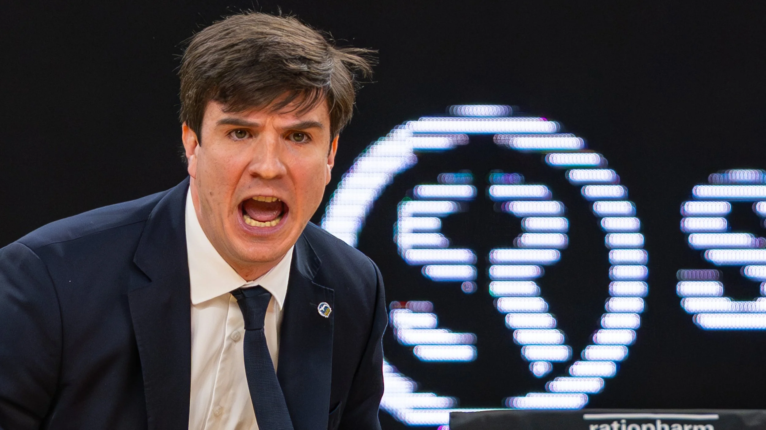 Ein Mann in einem dunklen Anzug schreit oder spricht energisch, im Hintergrund leuchtendes LED-Symbol eines Gehirns.