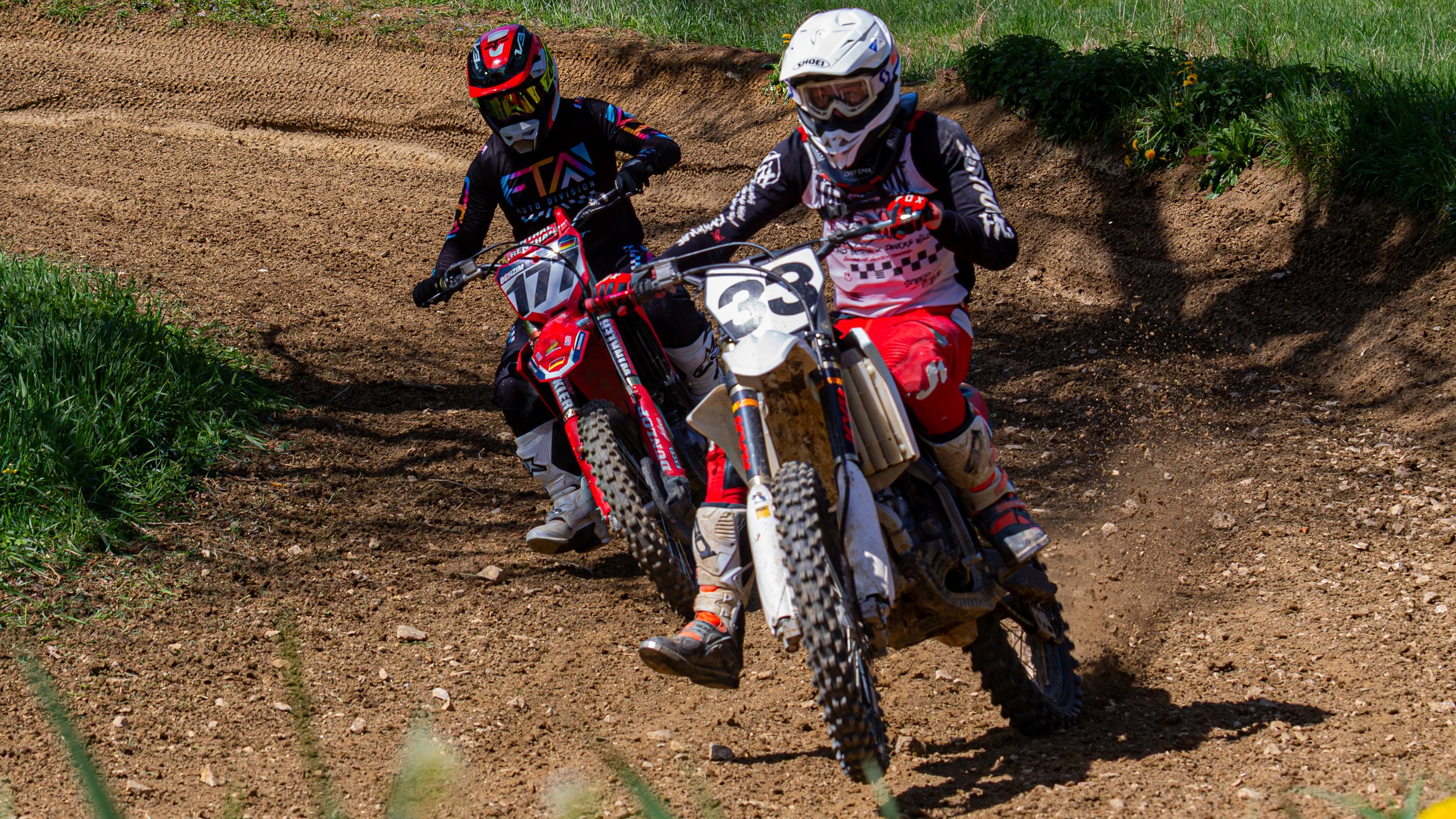 Zwei Motorradfahrer in Schutzkleidung und Helmen beim Kurvenfahren auf einem Dirttrack. Einer trägt ein schwarz-blau-rotes Outfit, der andere ein weiß-schwarz-rotes Outfit. Es ist ein sonniger Tag mit grüner Vegetation im Hintergrund.
