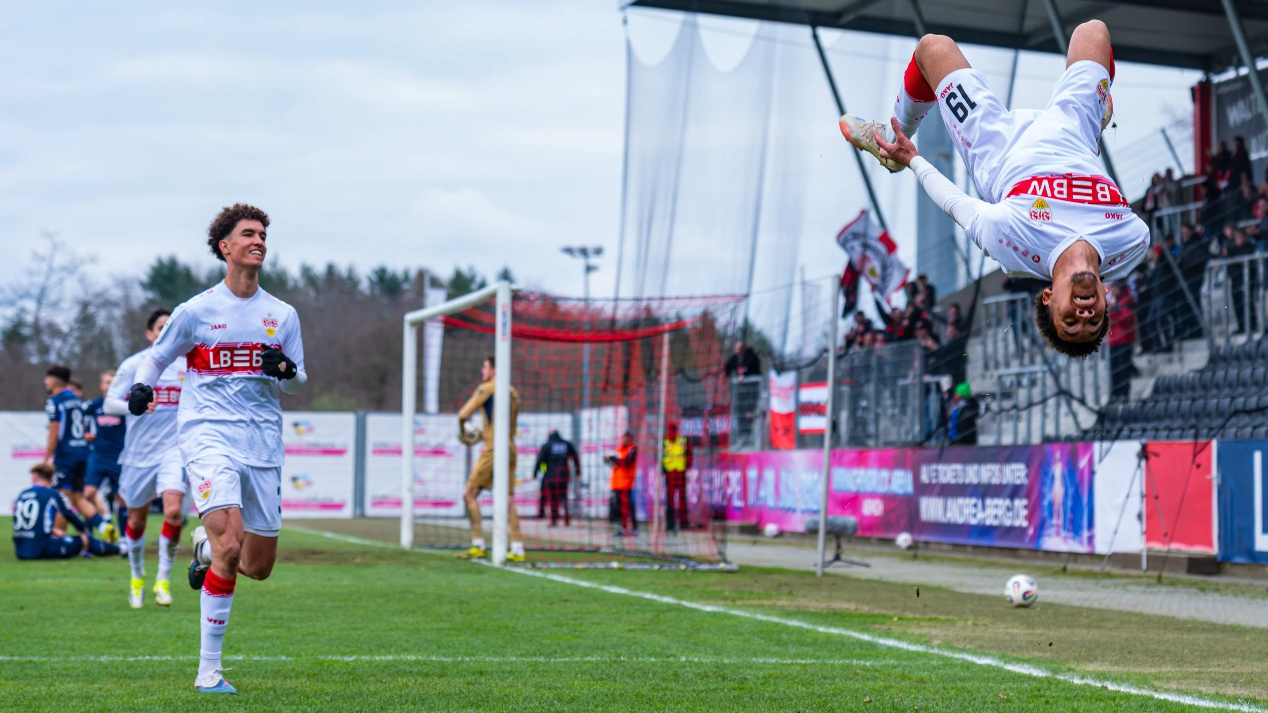 Fußballspieler jubeln nach einem Tor, einer springt kopfüber in die Luft vor Fans im Stadion.