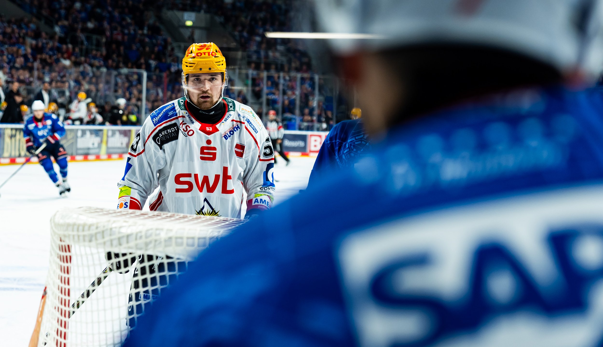 Ein Eishockeyspieler in weißem Trikot mit rotem Logo und gelber Helm steht vor dem Tor, während andere Spieler im Hintergrund auf dem Eis spielen.
