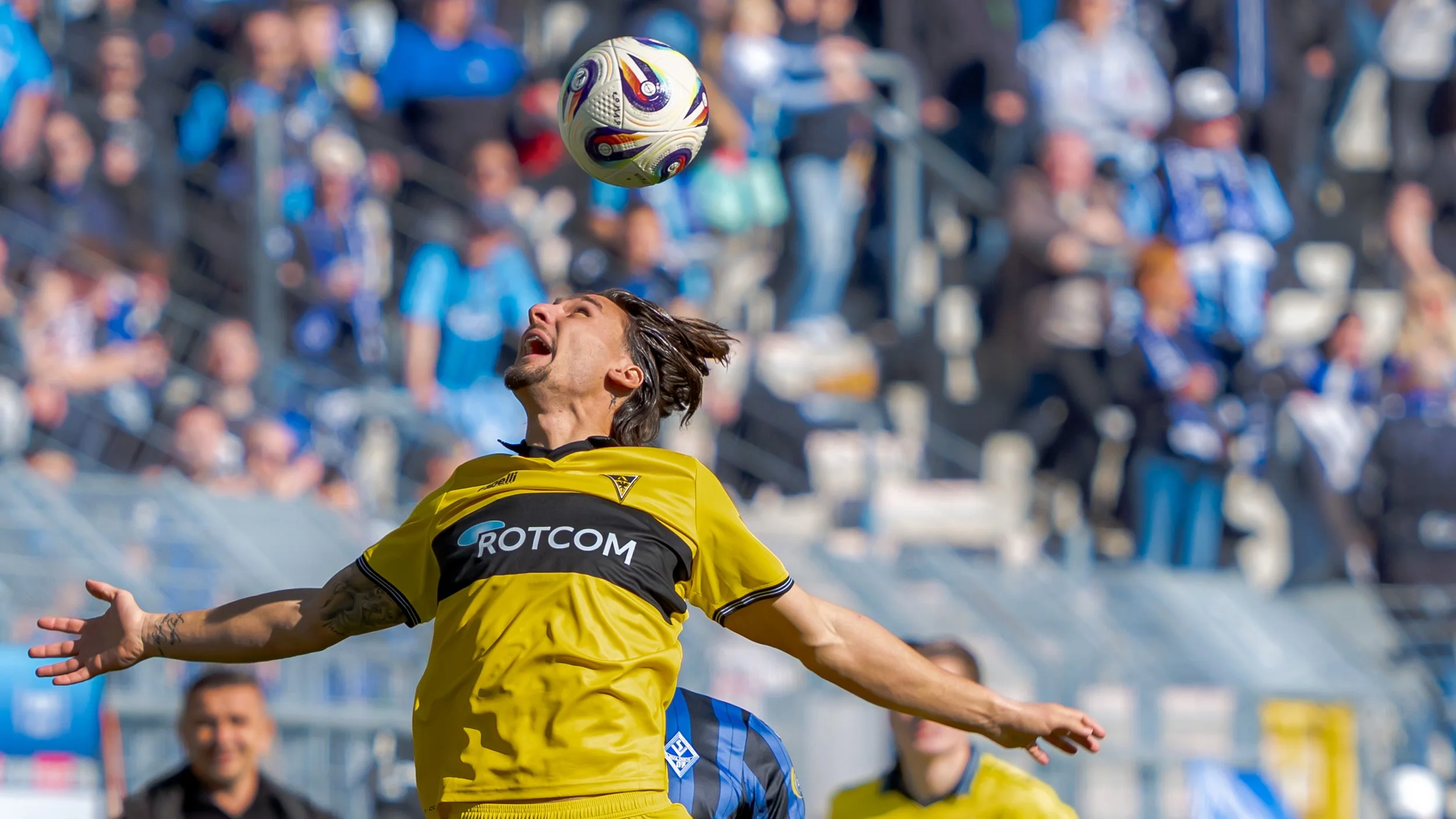Fußballspieler in gelbem Trikot springt für Kopfball in einem Spiel, im Hintergrund Fans im Stadion.