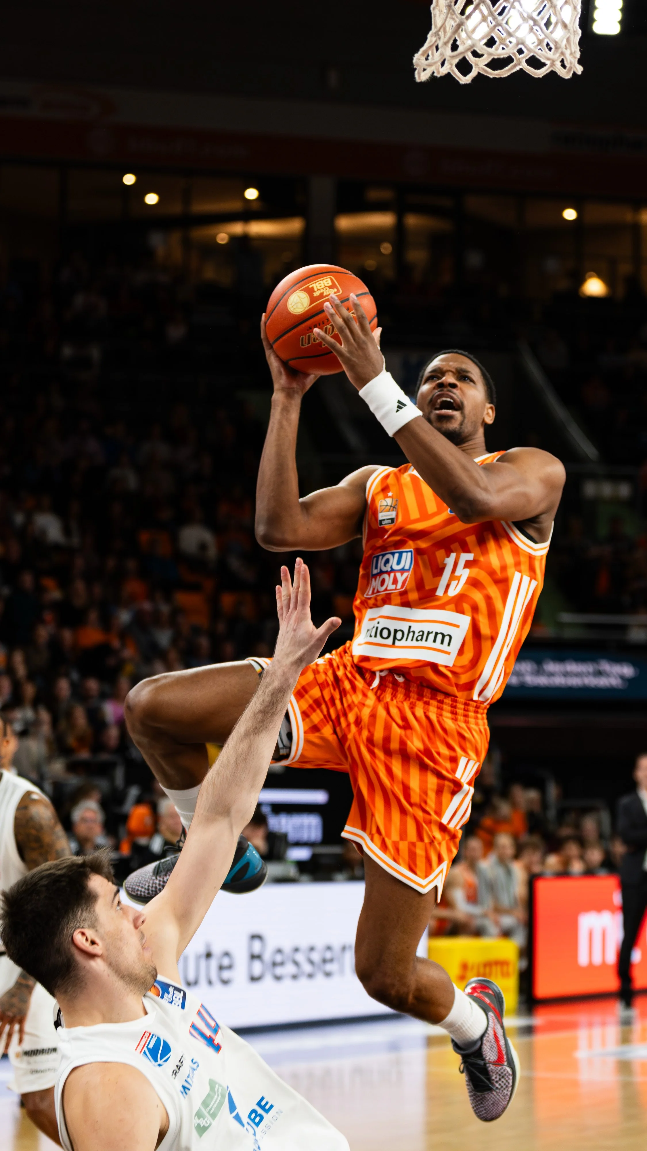 Ein Basketballspieler in orangefarbenem Trikot springt hoch, um den Ball während eines Spiels zu werfen, während der Verteidiger auf dem Boden liegt und versucht, den Wurf zu blockieren.