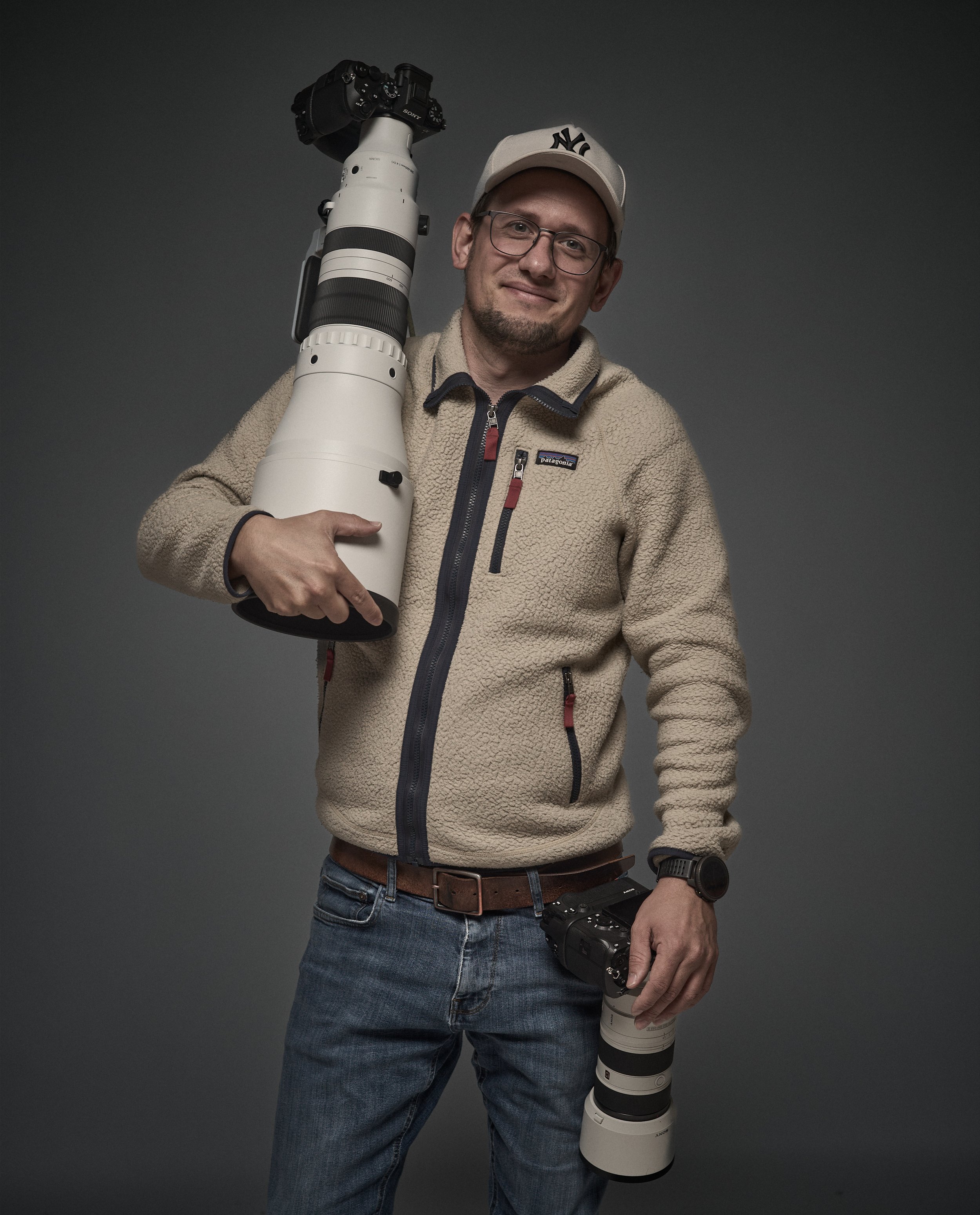 Mann trägt eine beige Fleecejacke, eine weiße Baseballmütze und besteht den Blick direkt in die Kamera, während er eine professionelle Kameraausrüstung mit einem großen Teleobjektiv trägt.