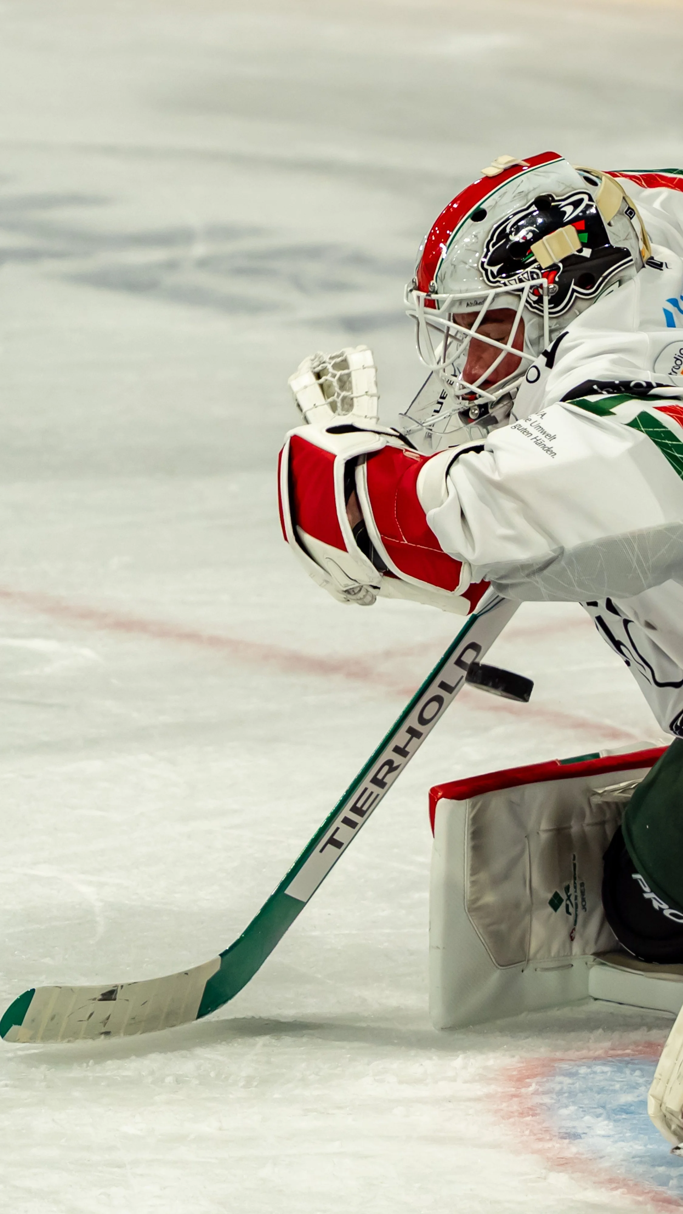 Ein Eishockeytorwart in Weiß mit rotem Helm und Handschuhen hält einen Puck auf dem Eis. Er ist in einer verschränkten Position, bereit zu stoppen.