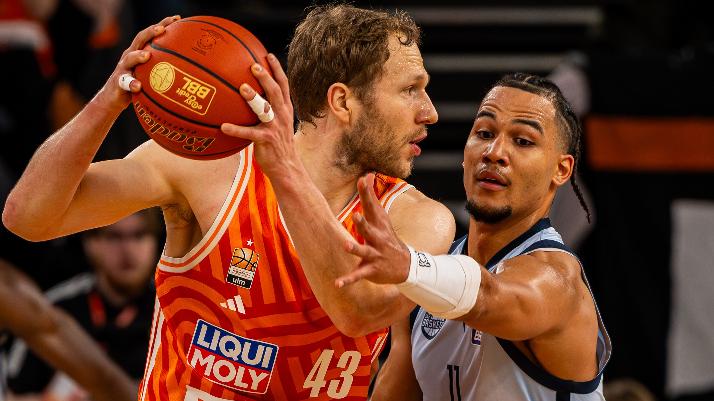 Zwei Basketballspieler im Kampf um den Ball während eines Spiels, einer trägt ein orangefarbenes Trikot und der andere ein weißes mit blauen Details.