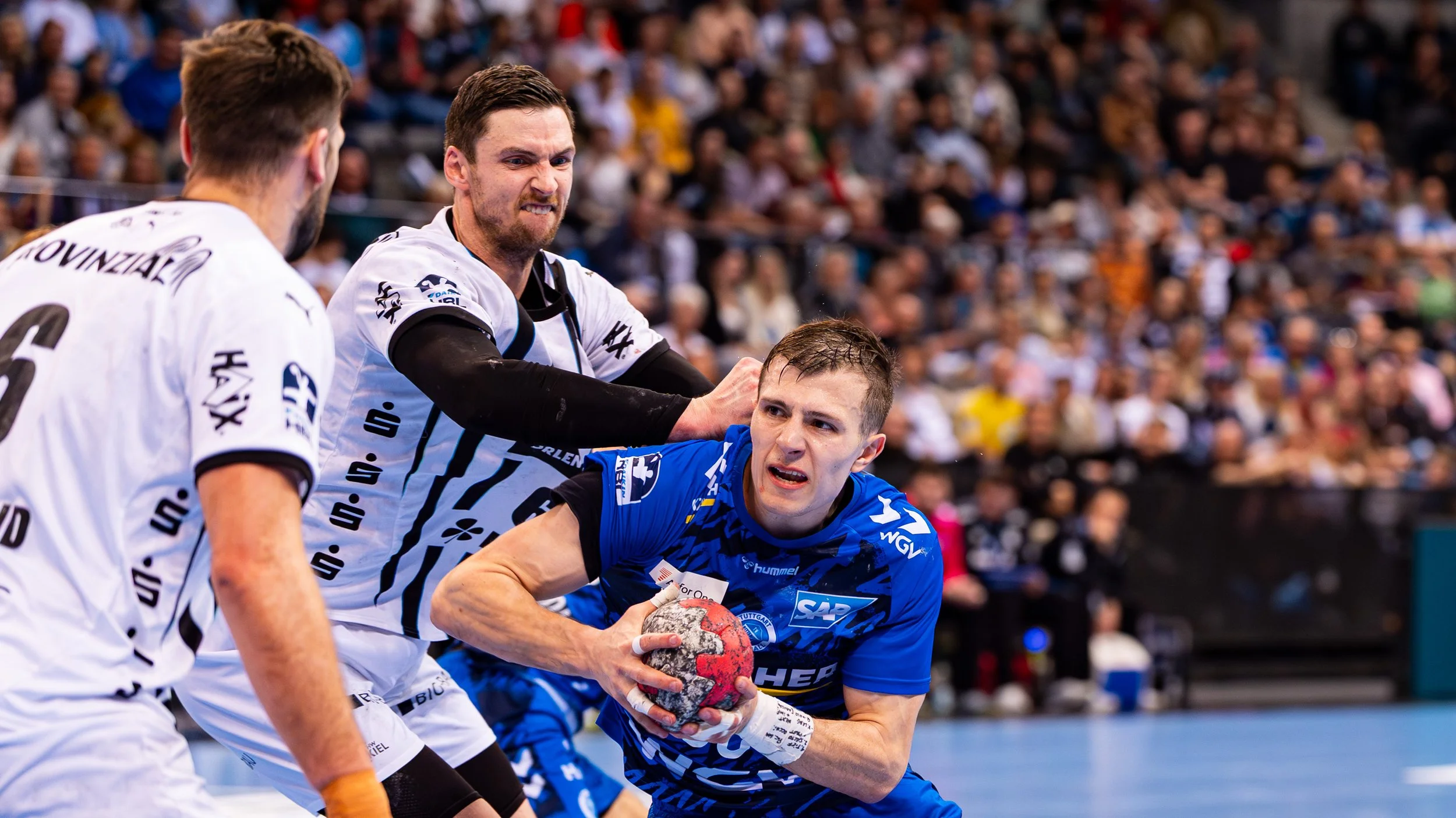 Handballspieler in einem Spiel, einer in blauer Kleidung hält den Ball, zwei in weißen Trikots versuchen ihn zu blockieren, im Hintergrund viele Zuschauer im Stadion.