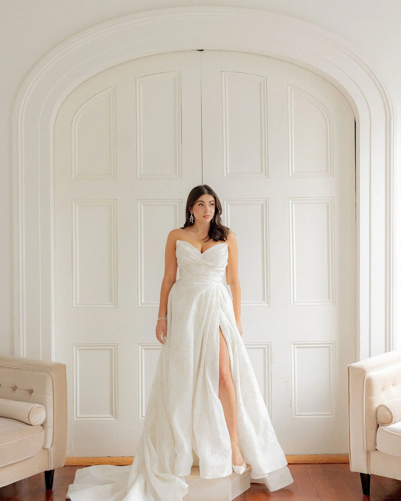 A woman in a white wedding dress with a slit in the skirt, standing in front of white paneled double doors in a bright room with wooden flooring.