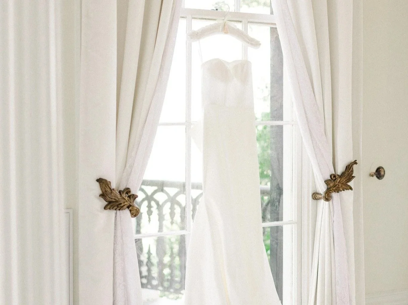 A wedding dress hanging on a window with cream curtains and antique brass curtain tiebacks, outside a balcony with a black railing and greenery.