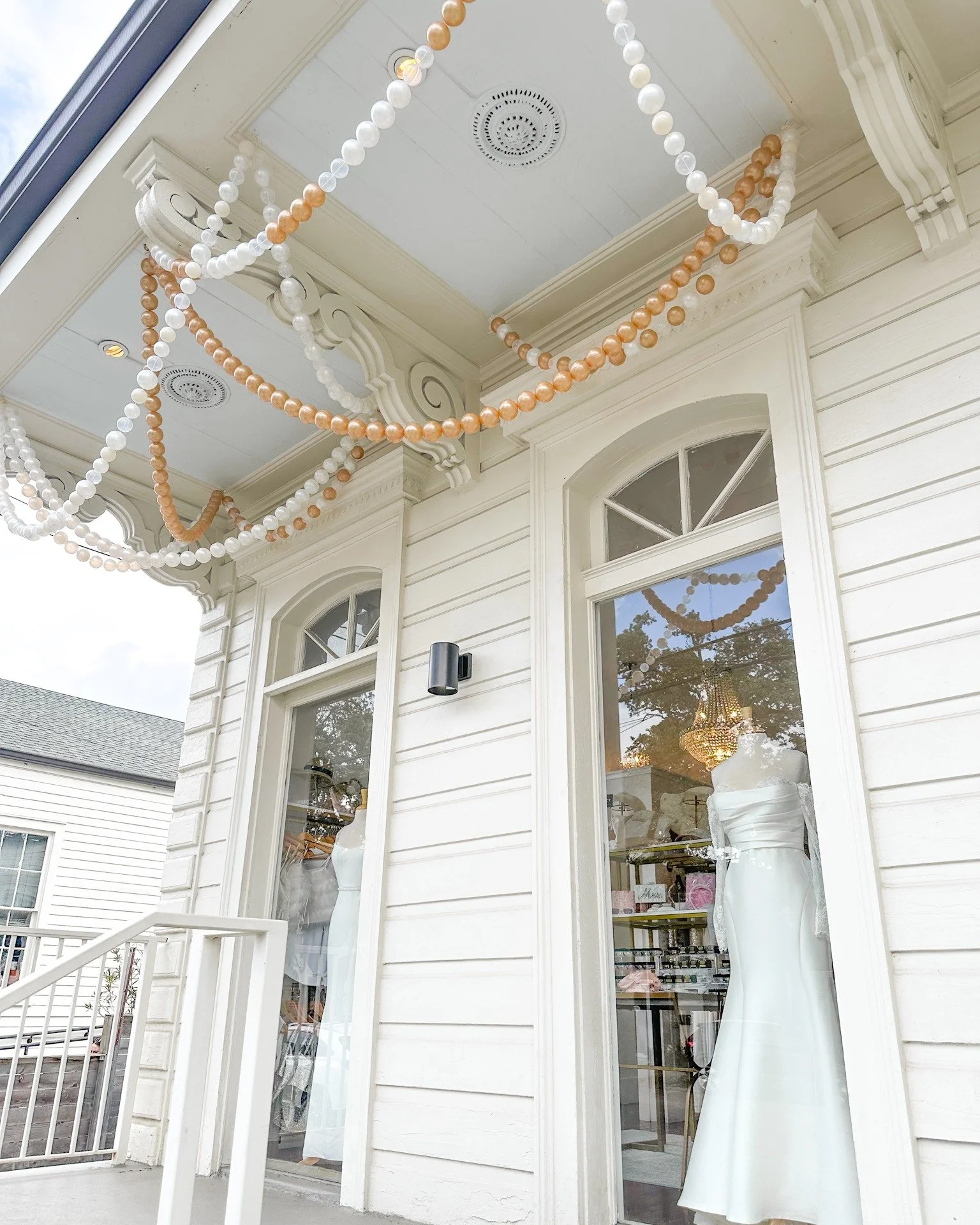 White building with large front windows featuring mannequins in white dresses, and decorated with pink and white bead garlands hanging from the porch ceiling.