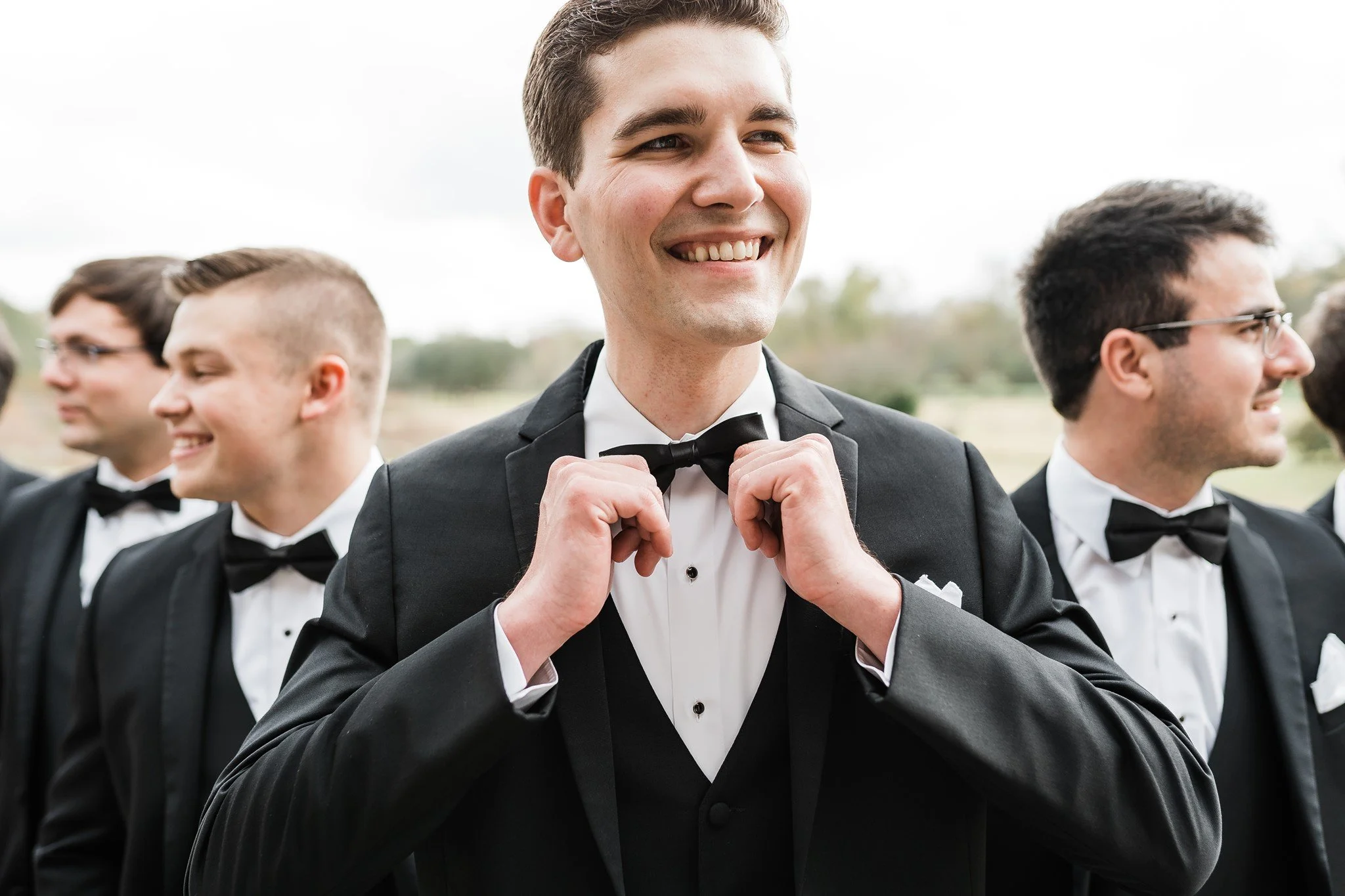A group of men dressed in formal tuxedos with bow ties, standing outdoors on a cloudy day. The central man is adjusting his bow tie and smiling while the others are talking or looking away.
