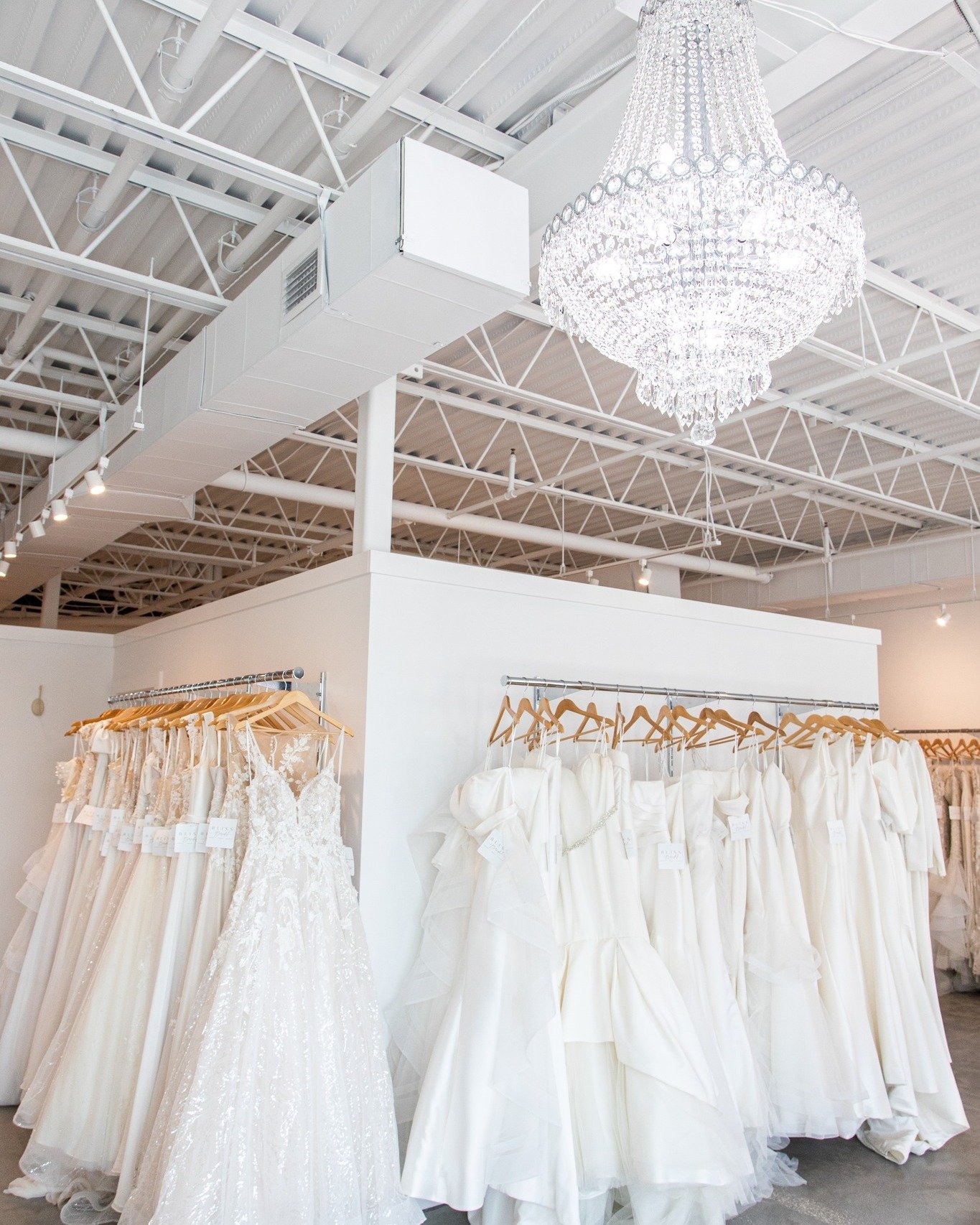 Wedding dresses on display in a bridal boutique, with white garments hanging on wooden hangers under a large crystal chandelier.