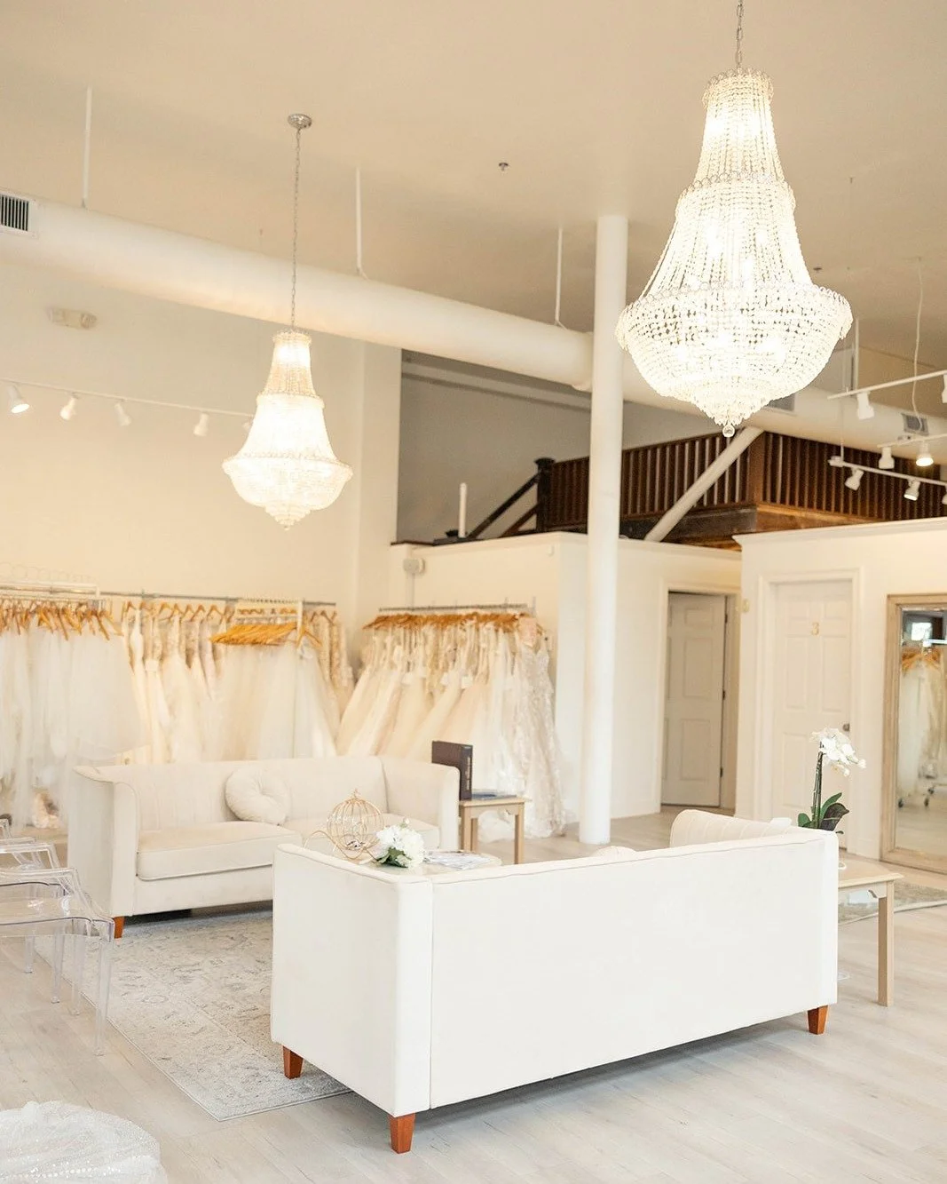 Interior of a boutique with white sofas, wedding dresses on racks, chandeliers, and a mirrored wall.